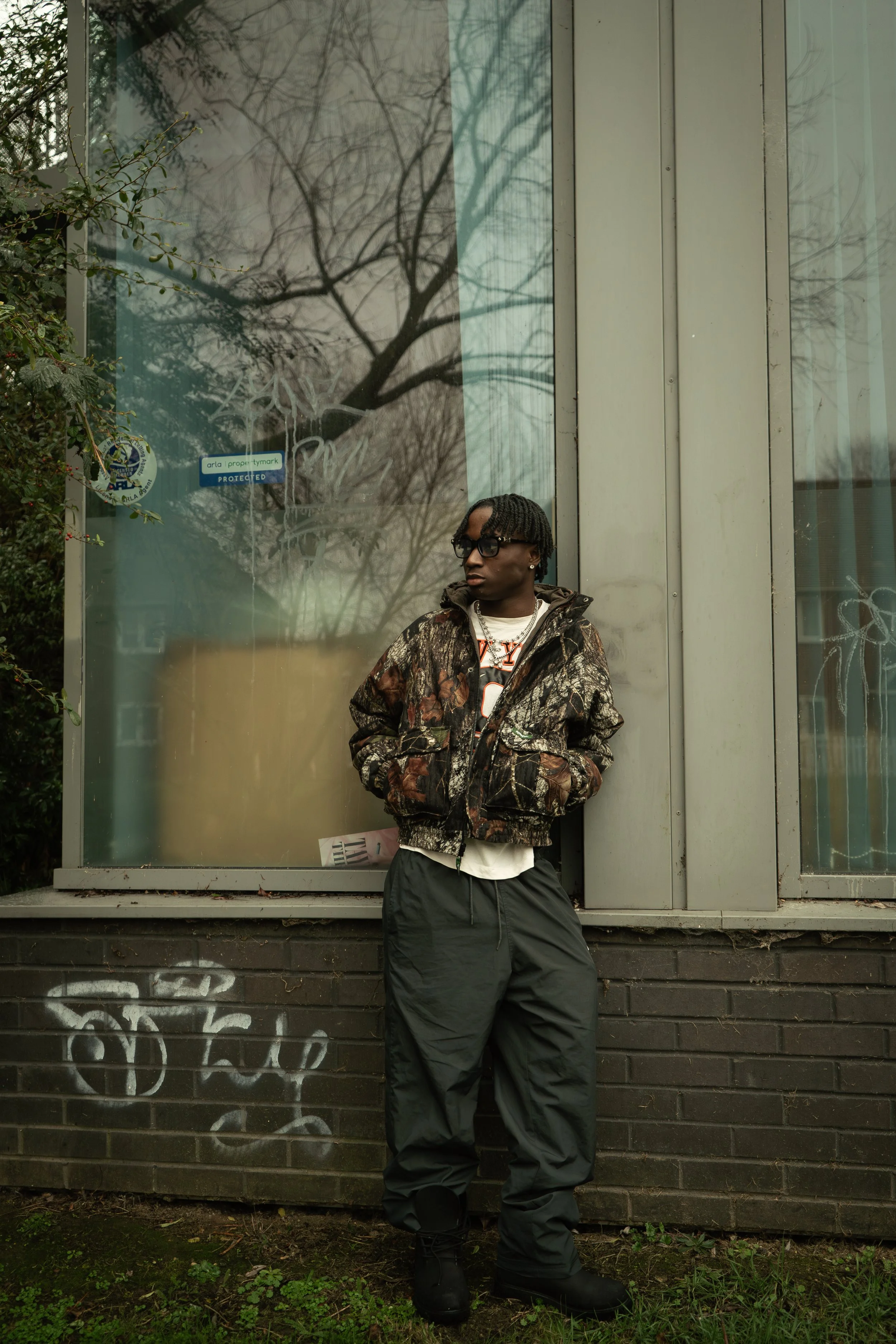 A person with dreadlocks, wearing glasses, a camouflage jacket, and black pants, standing in front of a glass window with tree reflections and graffiti on the lower brick wall.