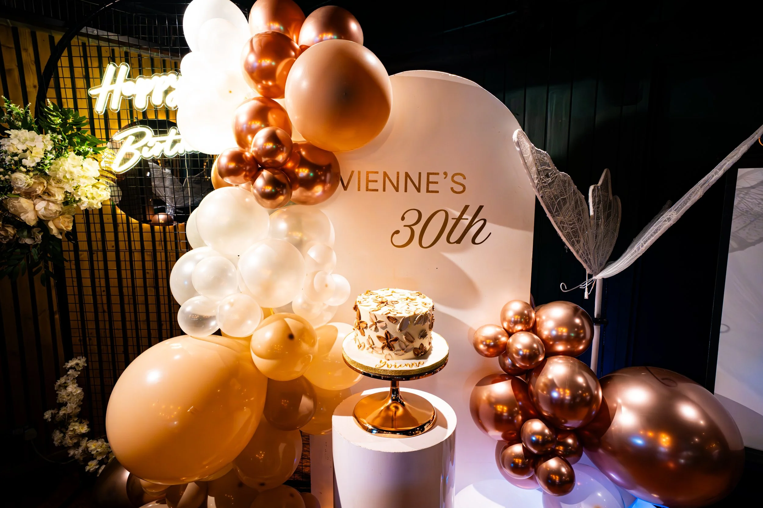 Decorative birthday celebration setup with a balloon arrangement in shades of white, gold, and rose gold, a cake with floral designs on a stand, a floral bouquet on the side, and a illuminated 'Happy Birthday' sign background.