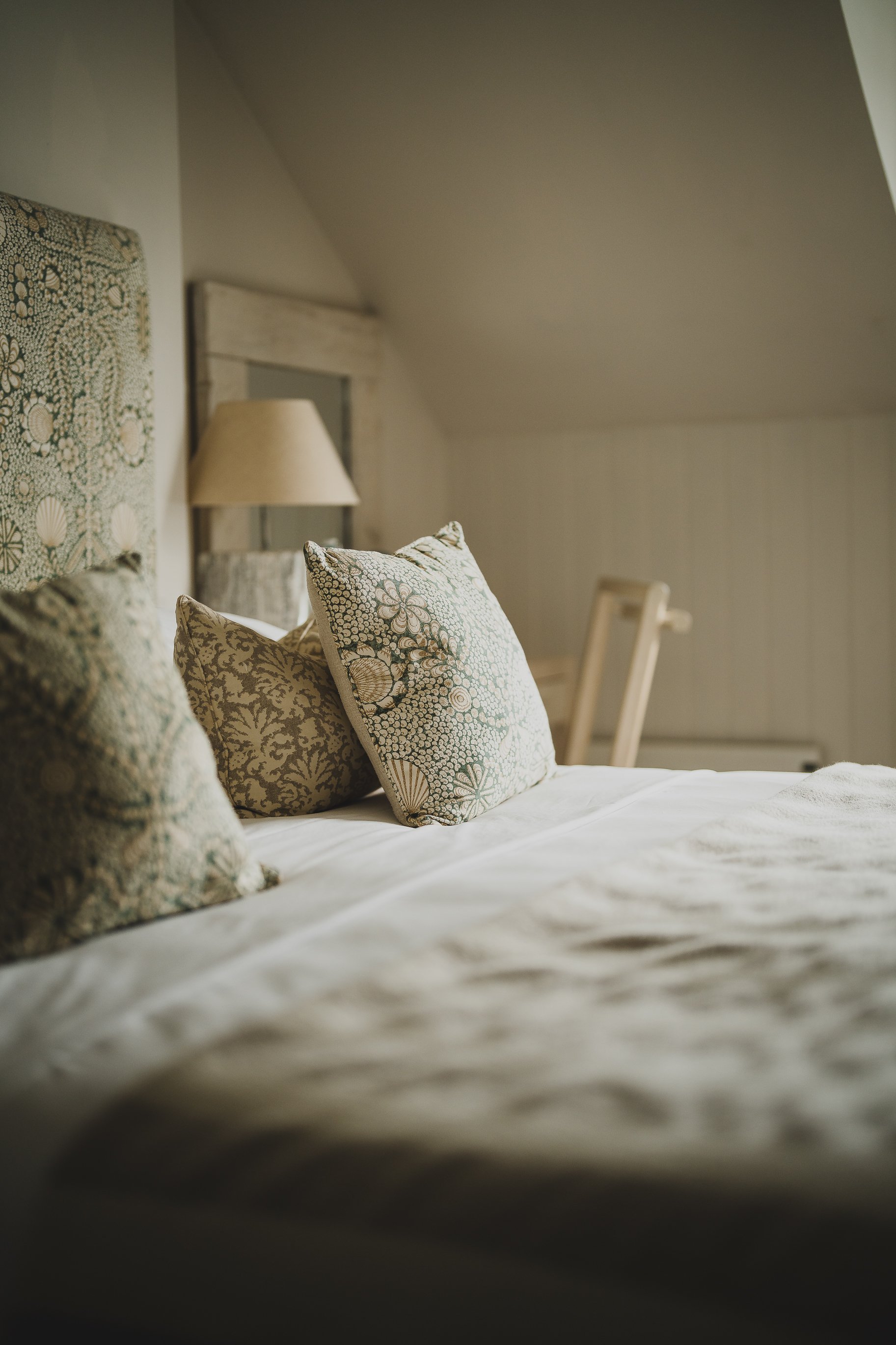 A cozy bedroom with a bed featuring European-style patterned bedding and pillows, a bedside lamp, and a chair in the background.