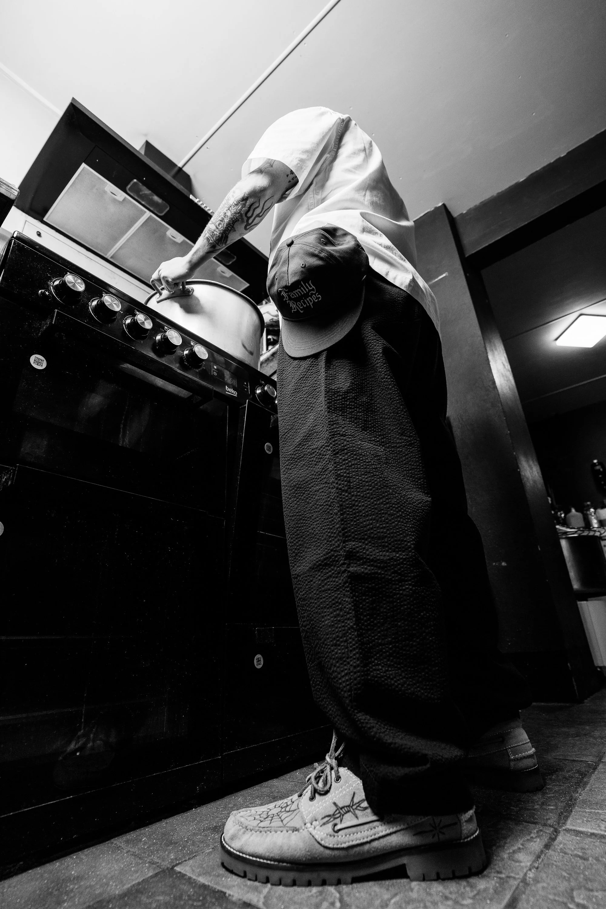 A person wearing a hat and sneakers is cooking on a stove in a kitchen, with the photo taken from a low angle in black and white.