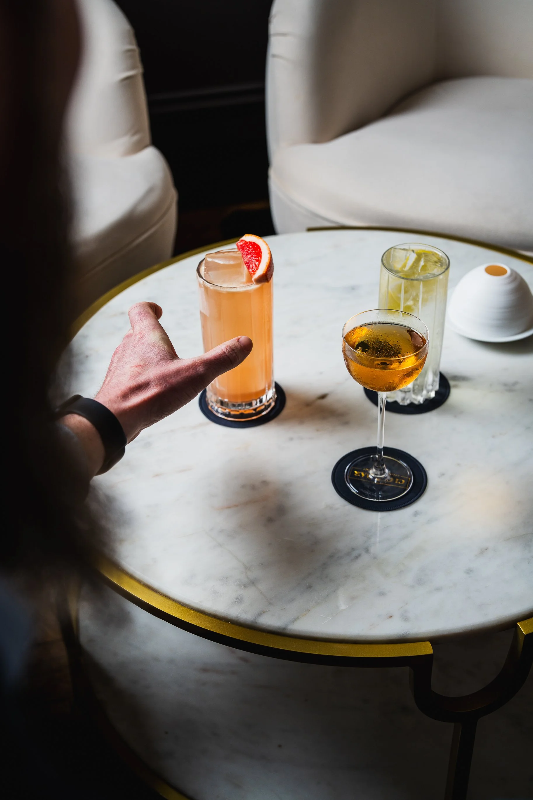 Person reaching for pink cocktail with grapefruit garnish on a white marble table with two other drinks and a white ceramic object.