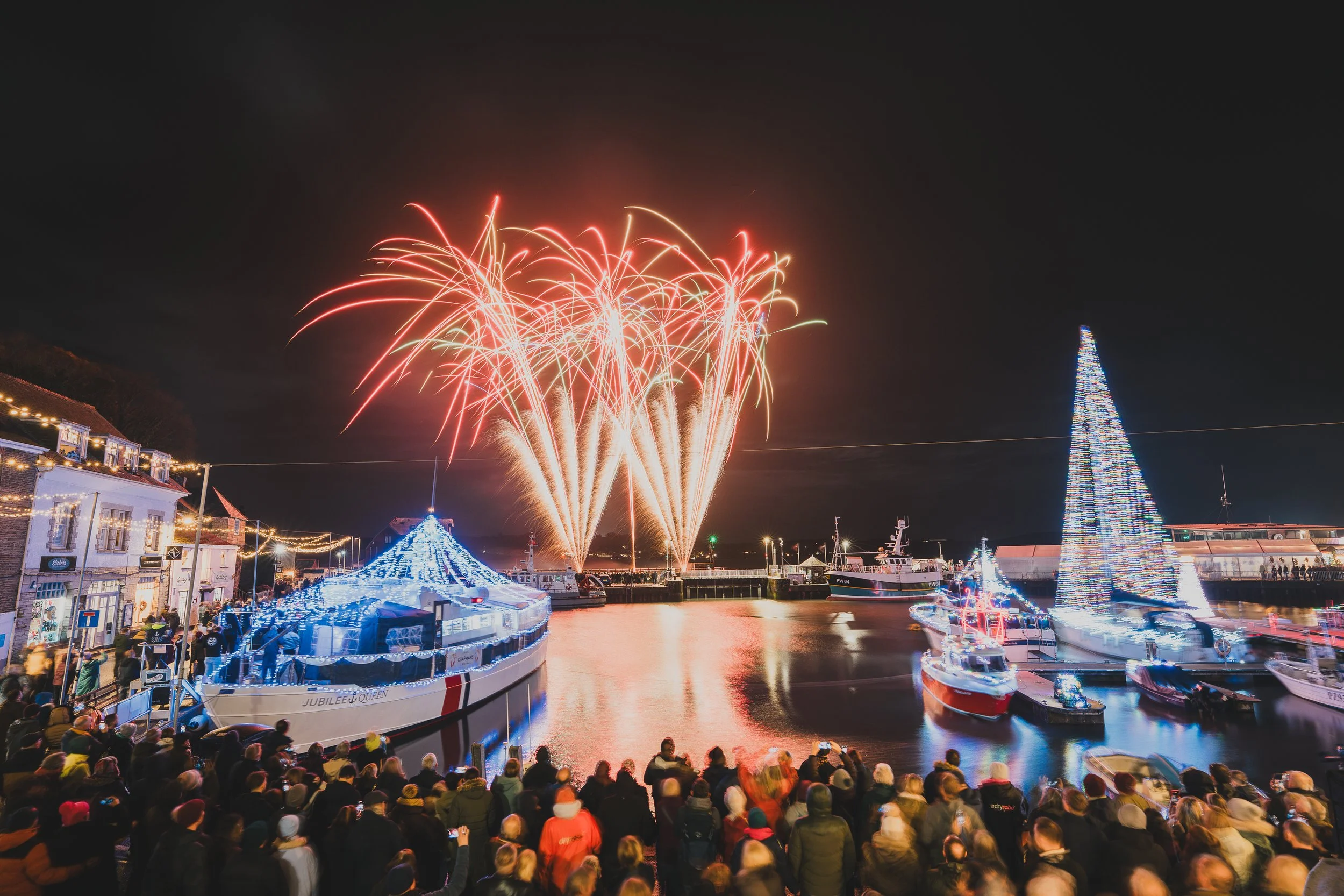 Nighttime harbor scene with boats decorated with Christmas lights, a large illuminated Christmas tree, and fireworks in the sky, with a crowd of people watching.