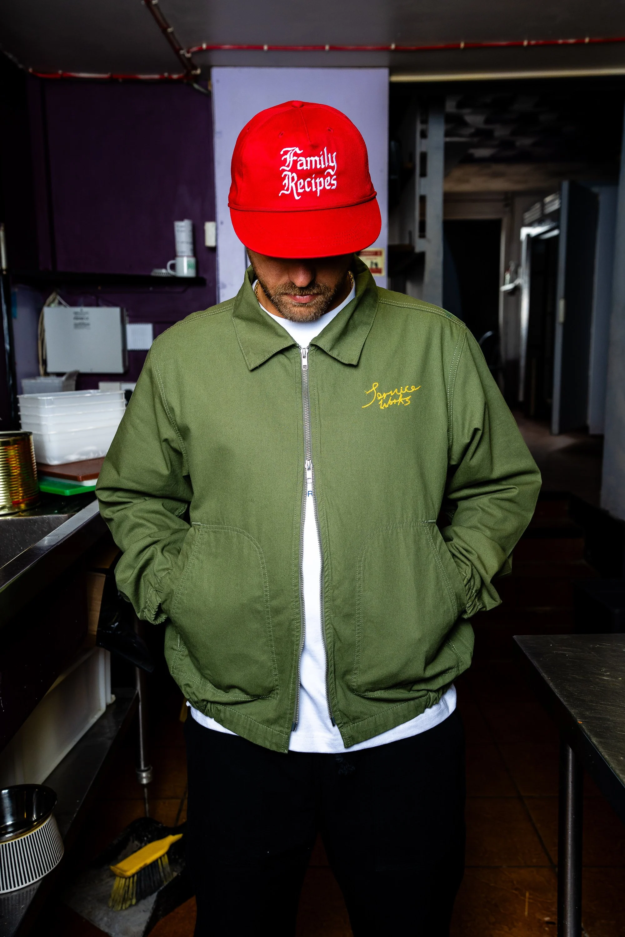 A man wearing a red cap with white embroidered text that reads 'Family Recipes' and a green jacket with yellow embroidered text that reads 'Sunrise Kitchen' inside a restaurant kitchen.
