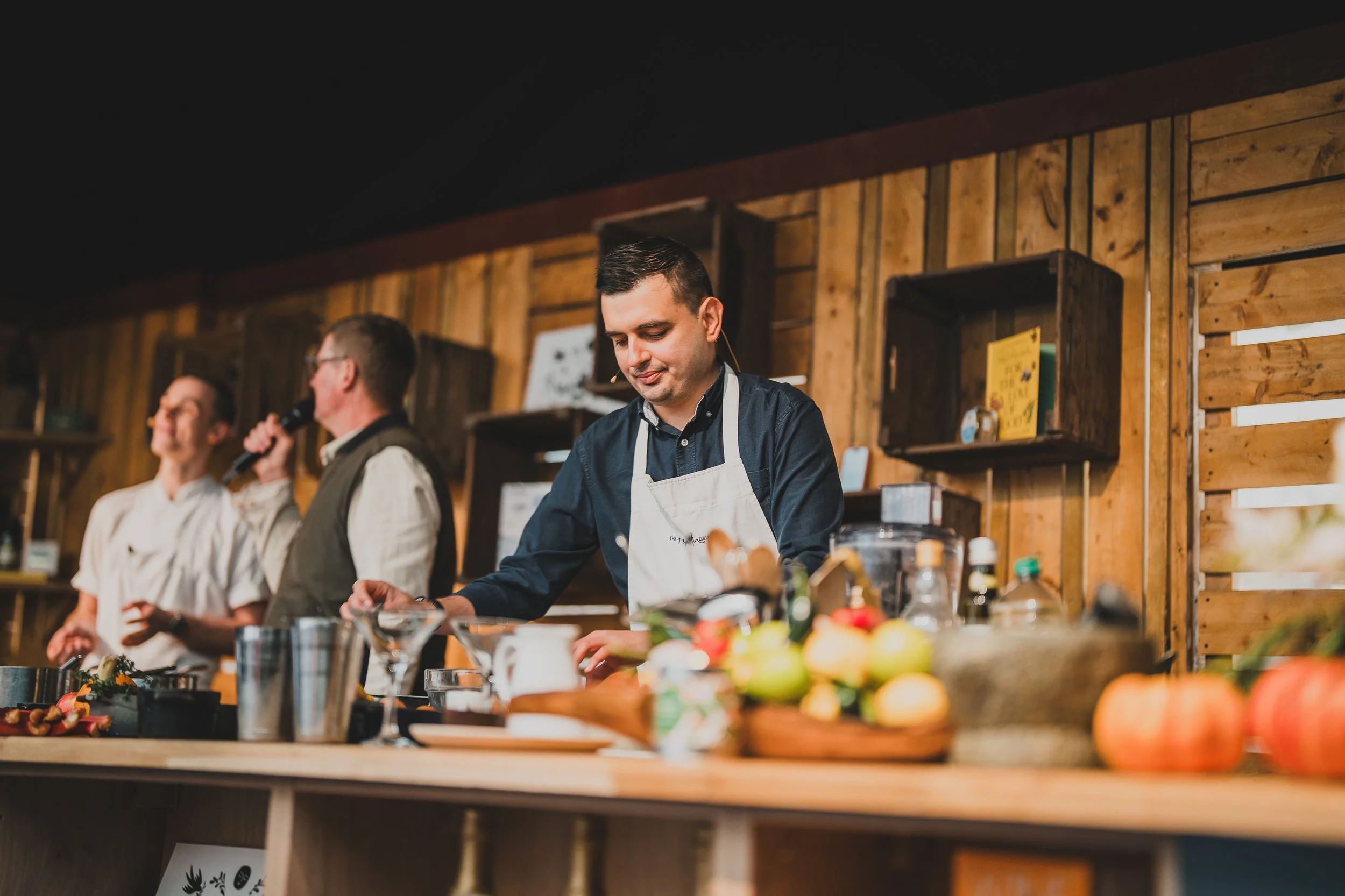 Three men in a kitchen or classroom setting with wood-paneled walls; the man in the foreground is wearing a white apron and is preparing food, while the two men in the background are speaking and smiling.