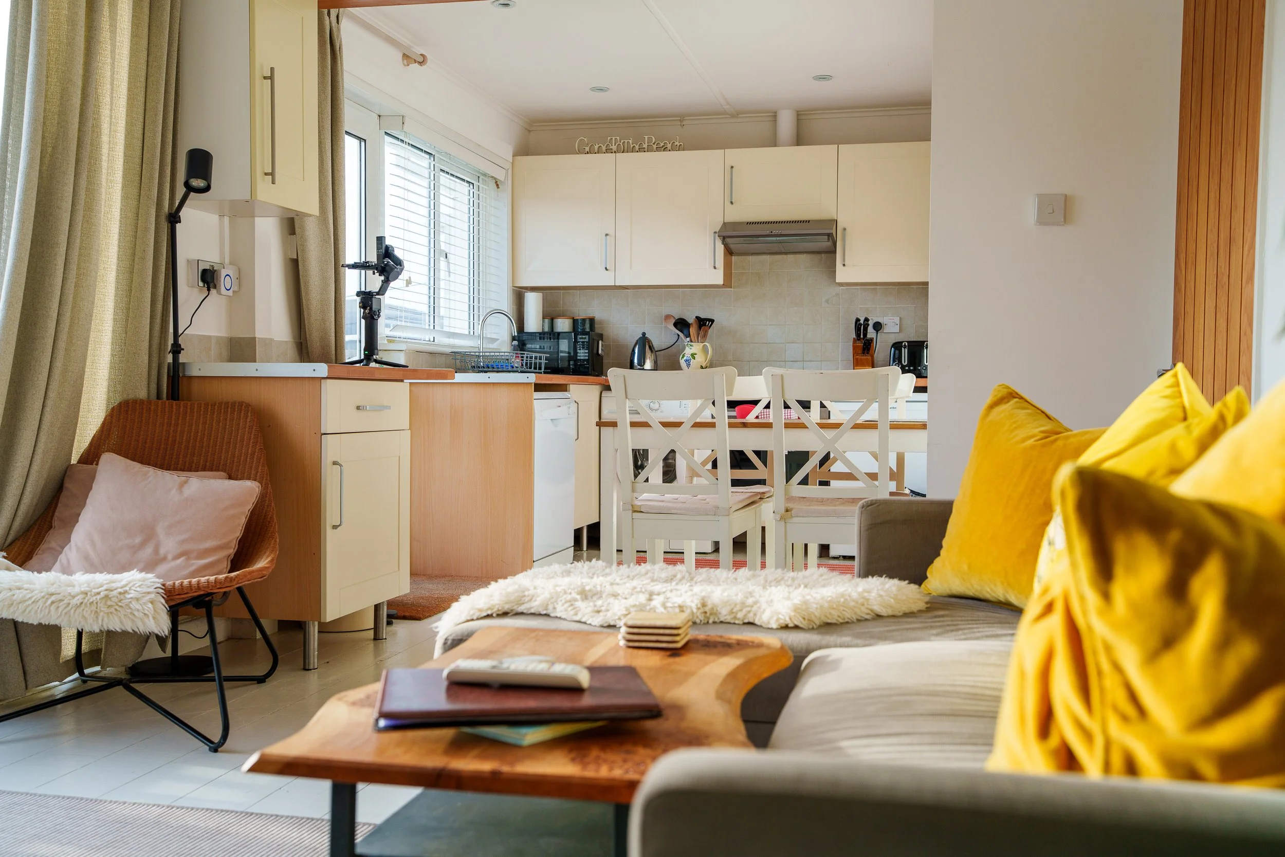 Living room with a beige sofa and yellow pillows, a wooden coffee table with a remote and folders, a cozy chair with a pillow and a blanket, a dining area with white chairs and a kitchen in the background featuring white cabinets, a window with blind