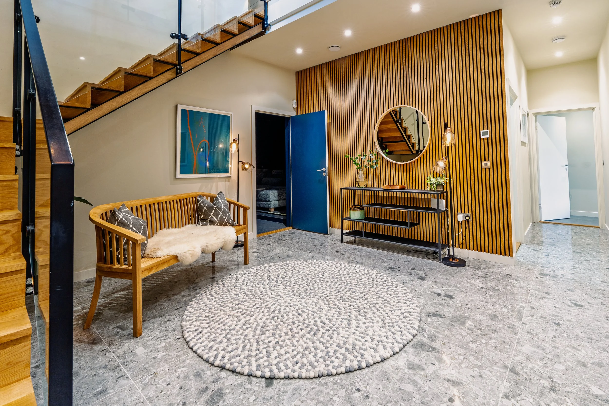 Modern living room entryway with wooden bench featuring pillows and a fur throw, a round rug on gray tile flooring, decorative wall with mirror, artwork, and vertical wooden slats, staircase, and doorway.
