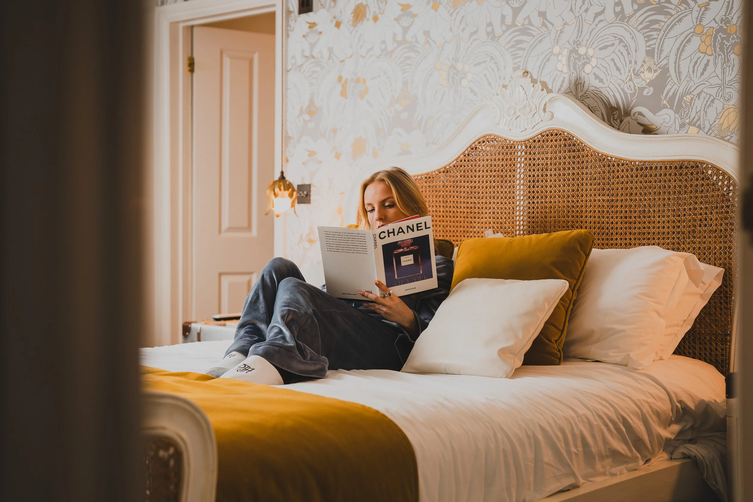 A woman is sitting on a bed in a cozy bedroom, reading a magazine titled 'Chanel'. The room features a decorative headboard, pillows, and floral wallpaper, with warm lighting from a bedside lamp.
