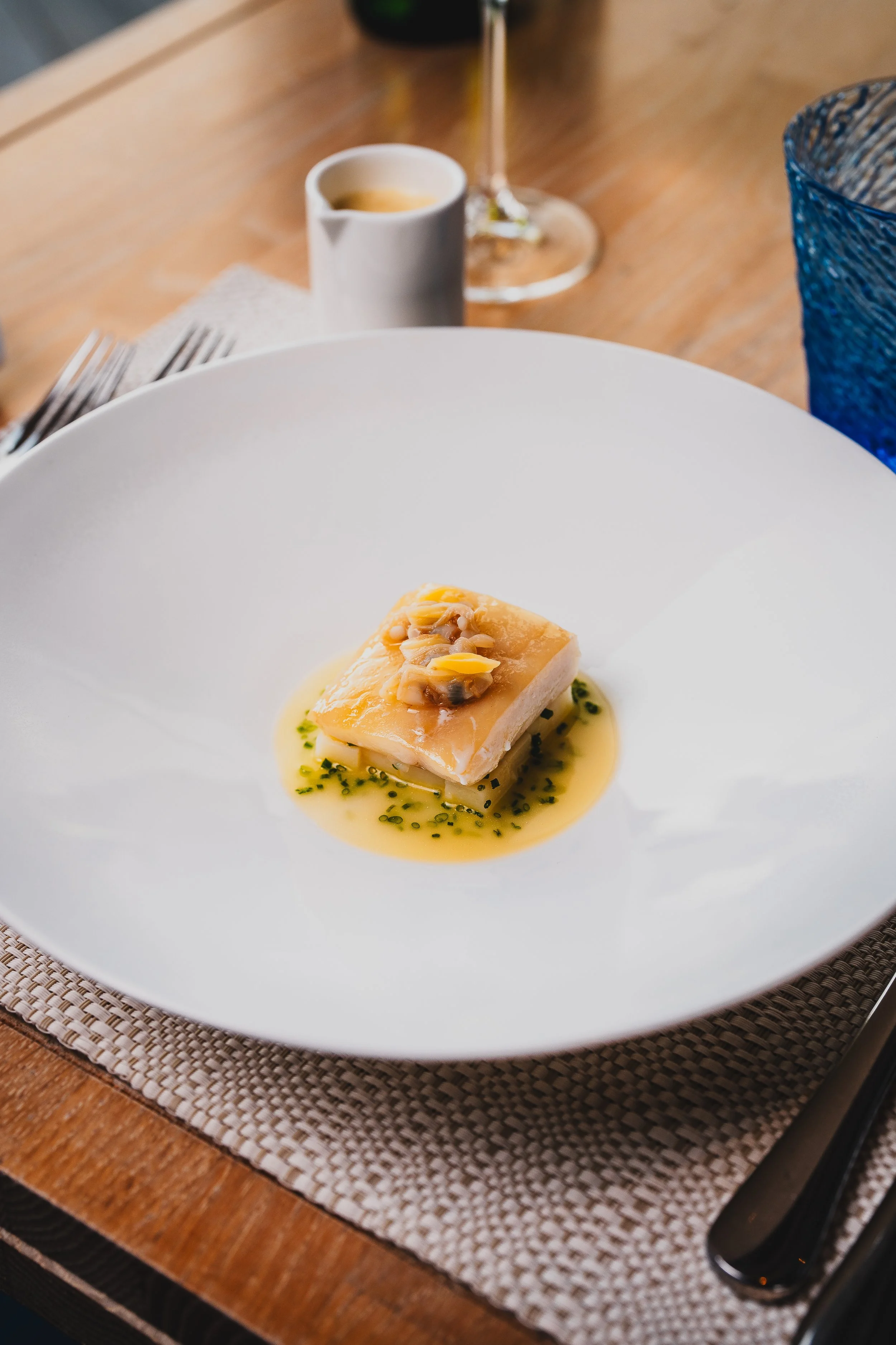 A plated dish with a small square piece of fish or seafood in light sauce, garnished with a small amount of seeds or nuts, and green herbs on a white plate.