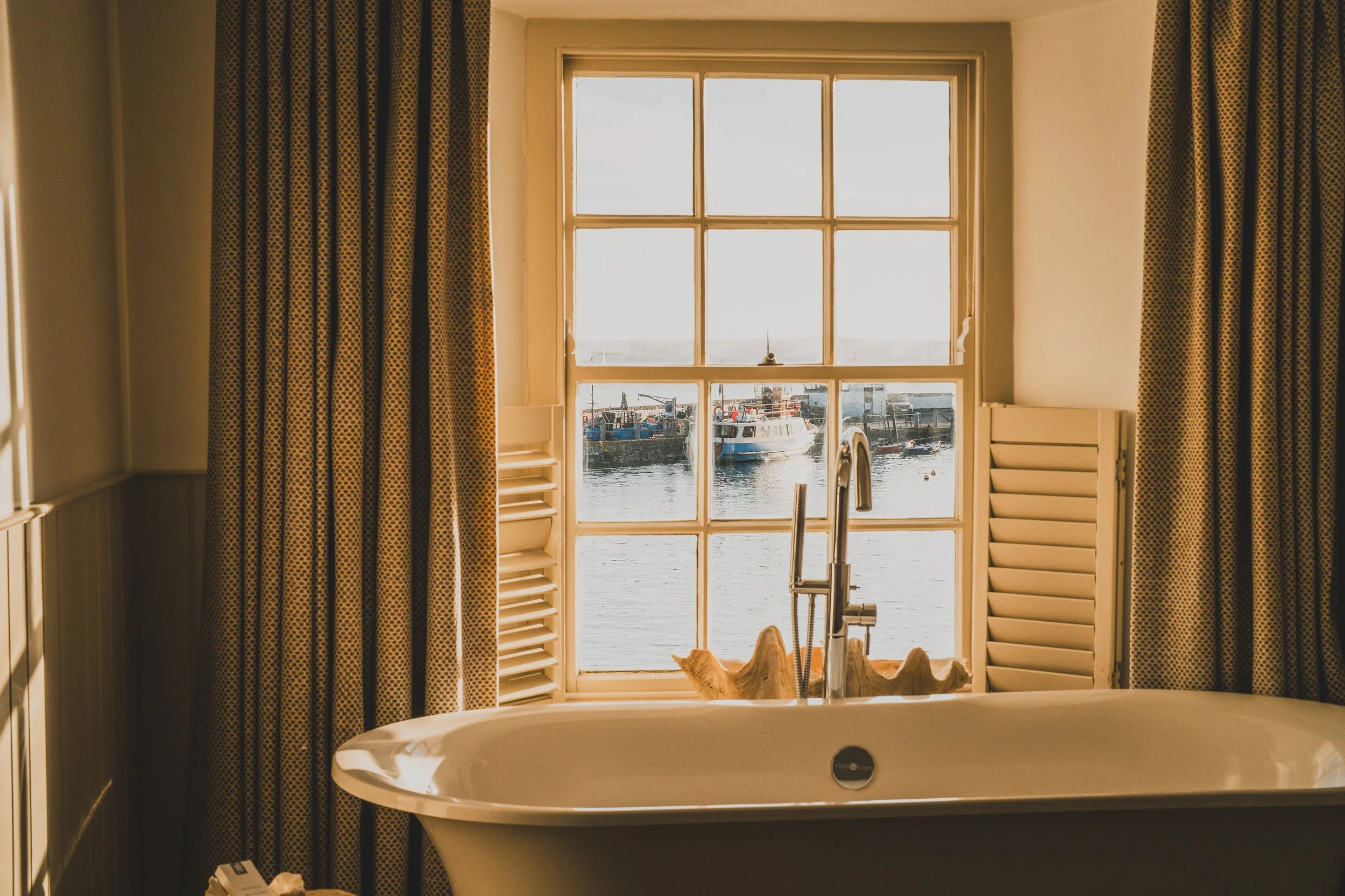 A bathroom with a bathtub looking out a window at a harbor with boats and water.