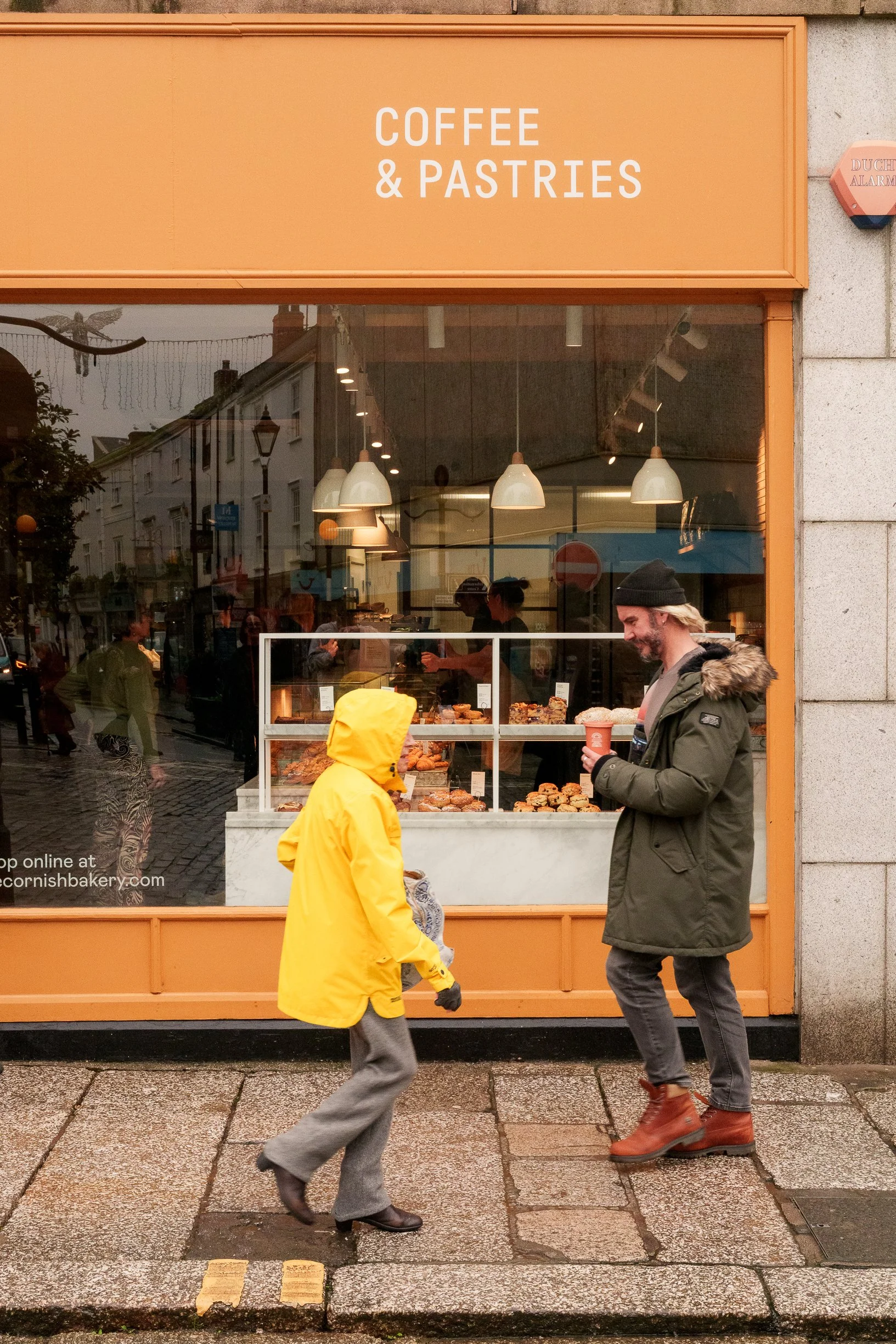 A storefront with a sign that reads 'Coffee & Pastries.' Two people are walking past the shop, one wearing a yellow raincoat and the other in a green coat with a black beanie, holding a coffee cup.