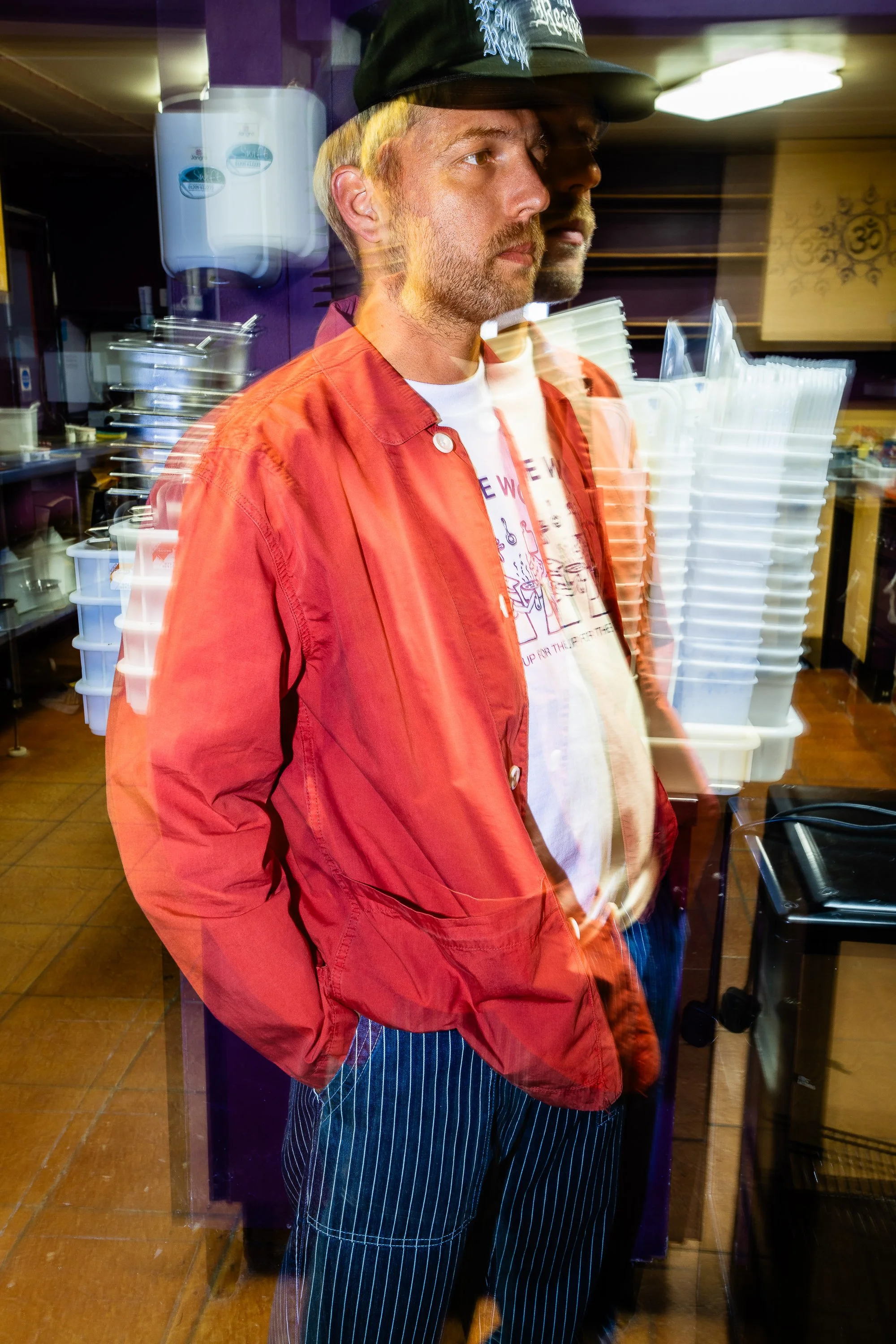 A man wearing a black cap, red jacket, and striped pants standing behind a glass window, creating a double exposure effect with a blurred outline of his face and body.