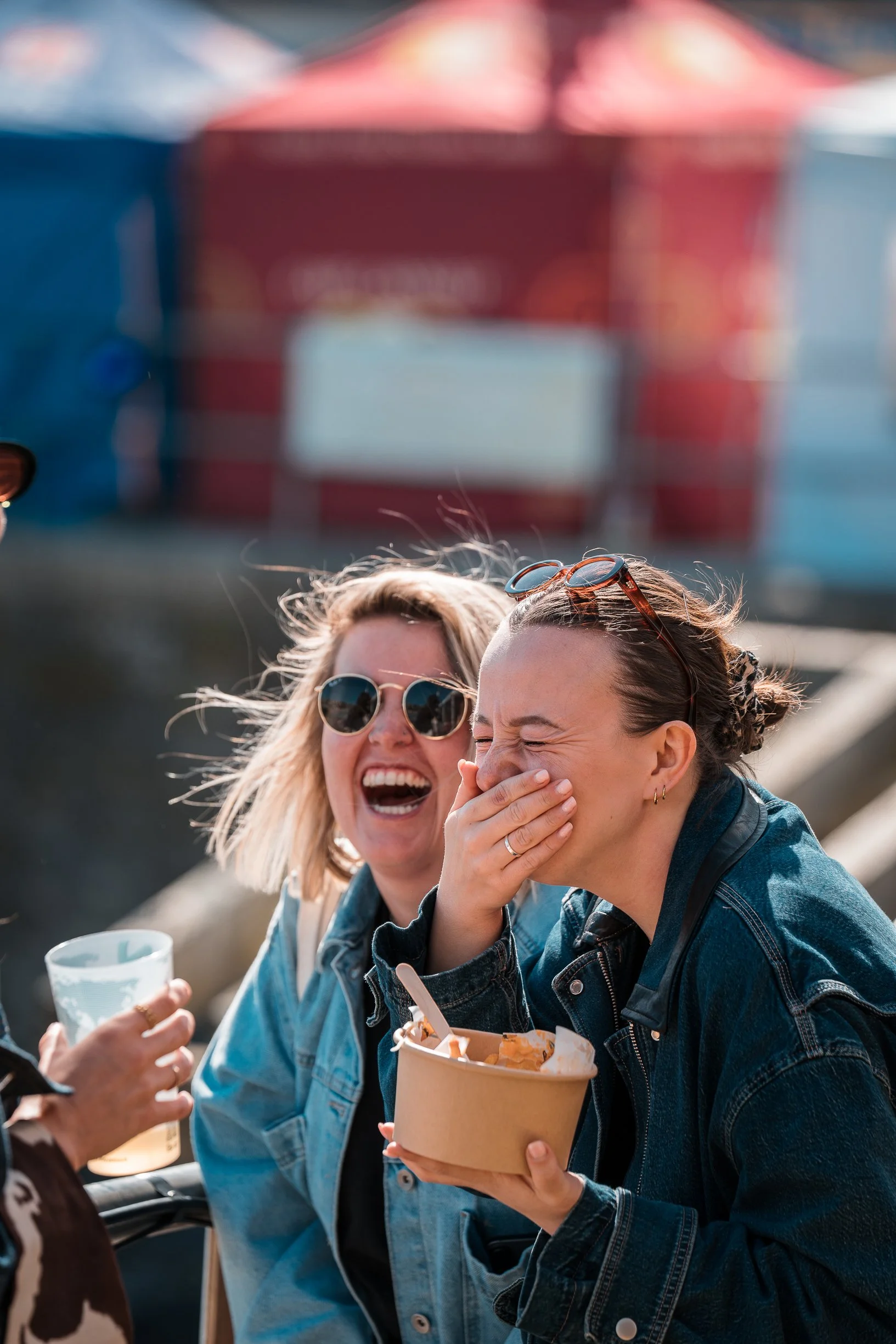 Two women laughing and talking outdoors, one holding a cup and the other holding food in a container, with a blurred red building in the background.
