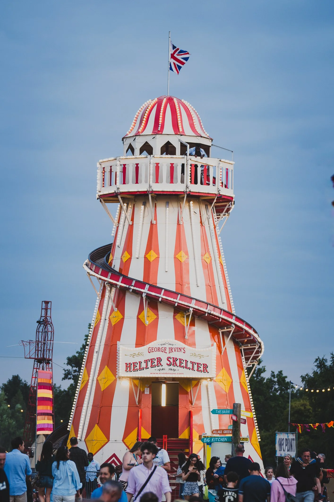 Crowd of people gathered in front of a colorful amusement park ride called the Helter Skelter at a fair, with a British flag flying at the top.