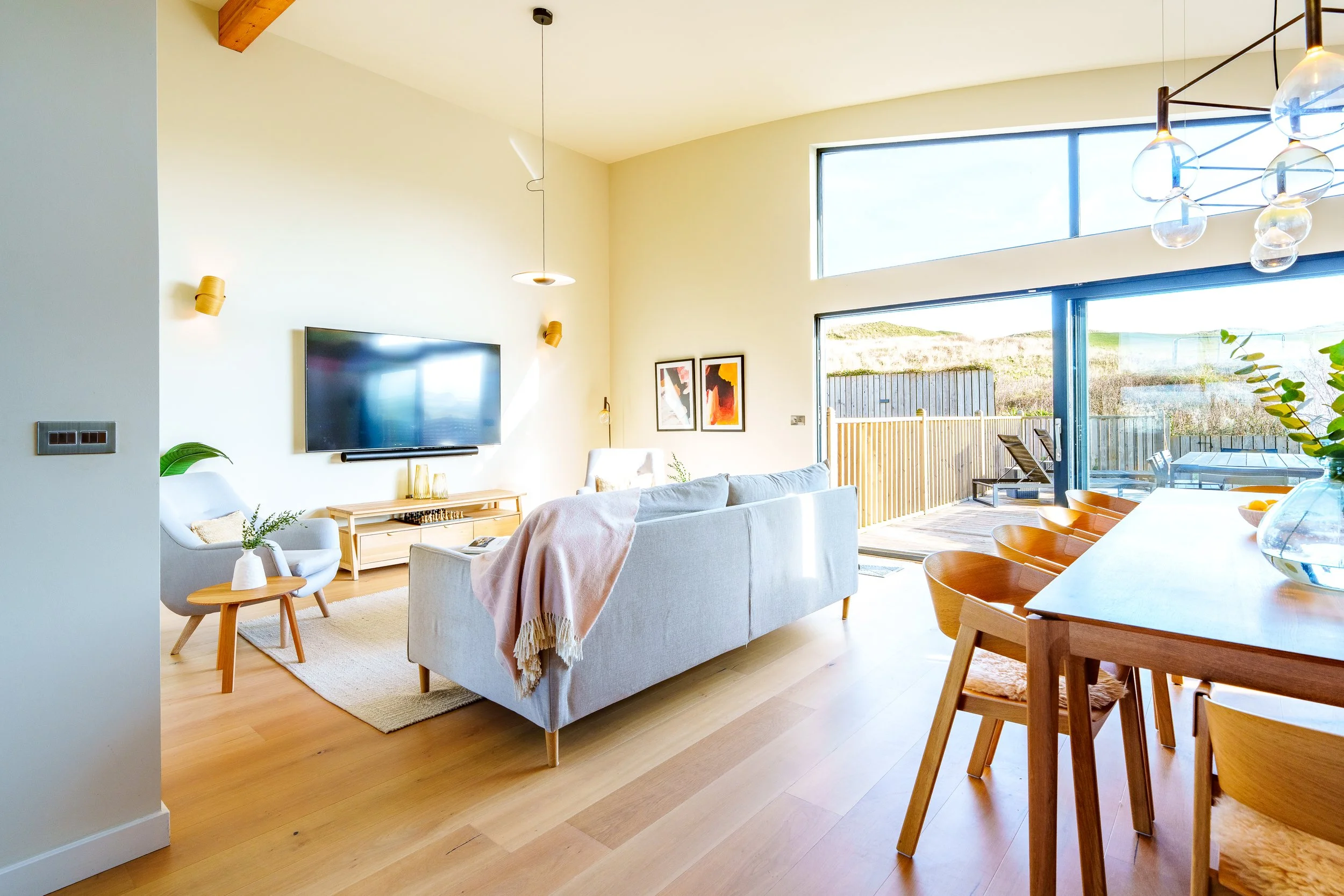 Bright living room with large sliding glass door leading to a wooden deck, light-colored sofa, armchair, wooden dining table with chairs, and modern pendant lights.