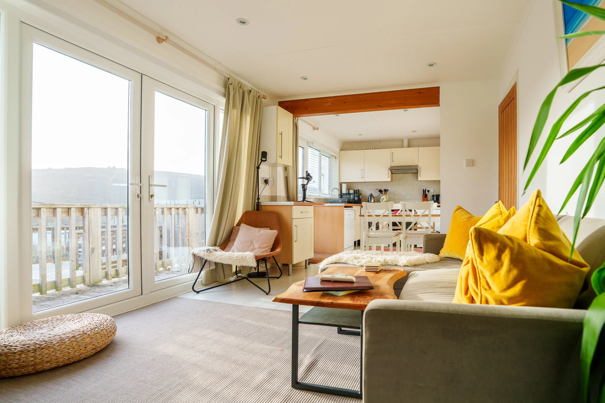Bright living room with large glass doors leading to a balcony, beige curtains, a beige sofa with yellow pillows, a wooden coffee table, a pink chair, and an open kitchen in the background with white cabinets and dining table.