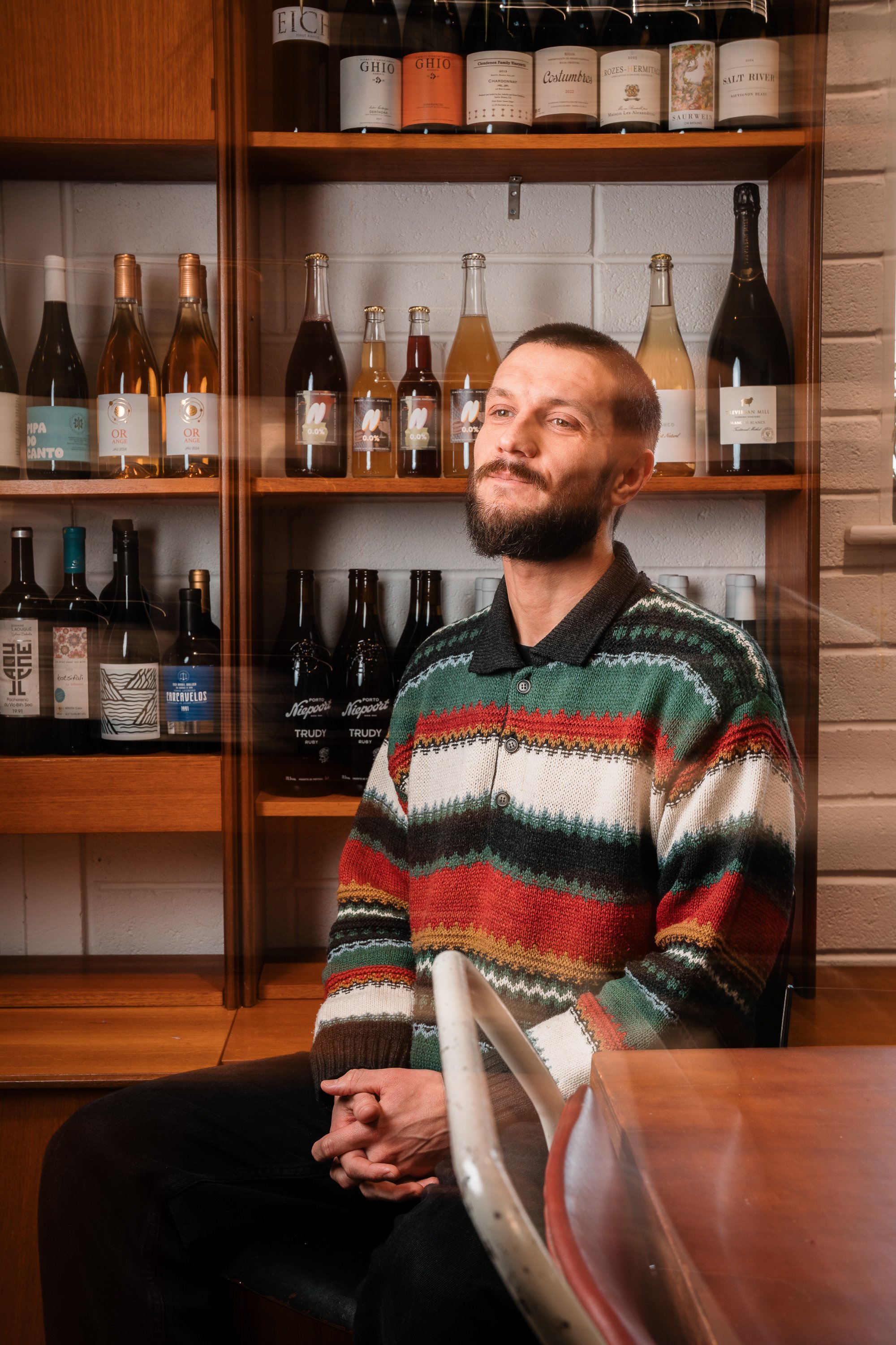 A man with a beard and short hair, wearing a colorful striped sweater, is seated with clasped hands, in front of a glass barrier, in a room with shelves of various wine bottles and liquor bottles against a brick wall.