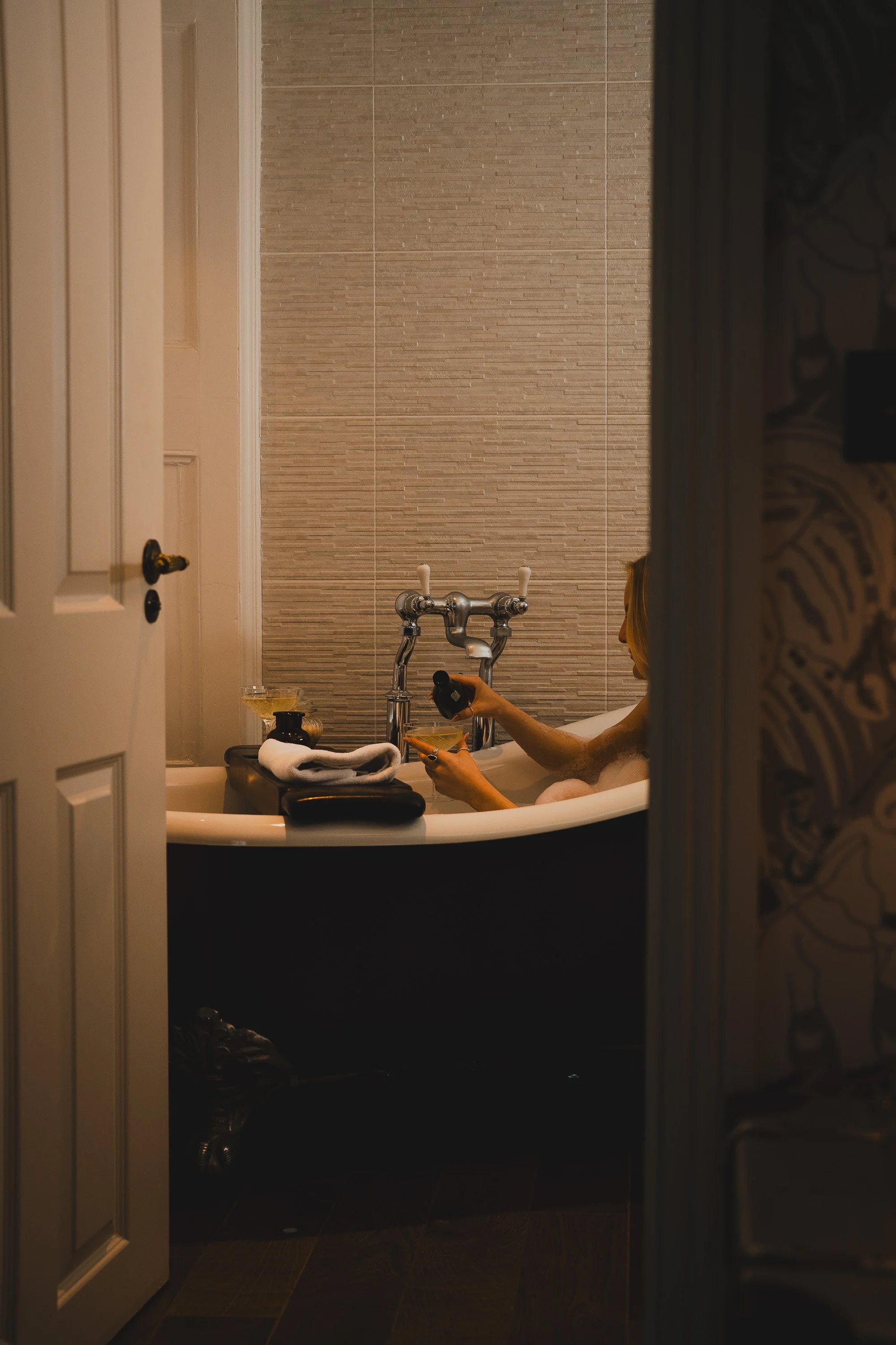 Person in a bathtub pouring a drink, with bath essentials on a tray, in a bathroom with modern wall tiles.