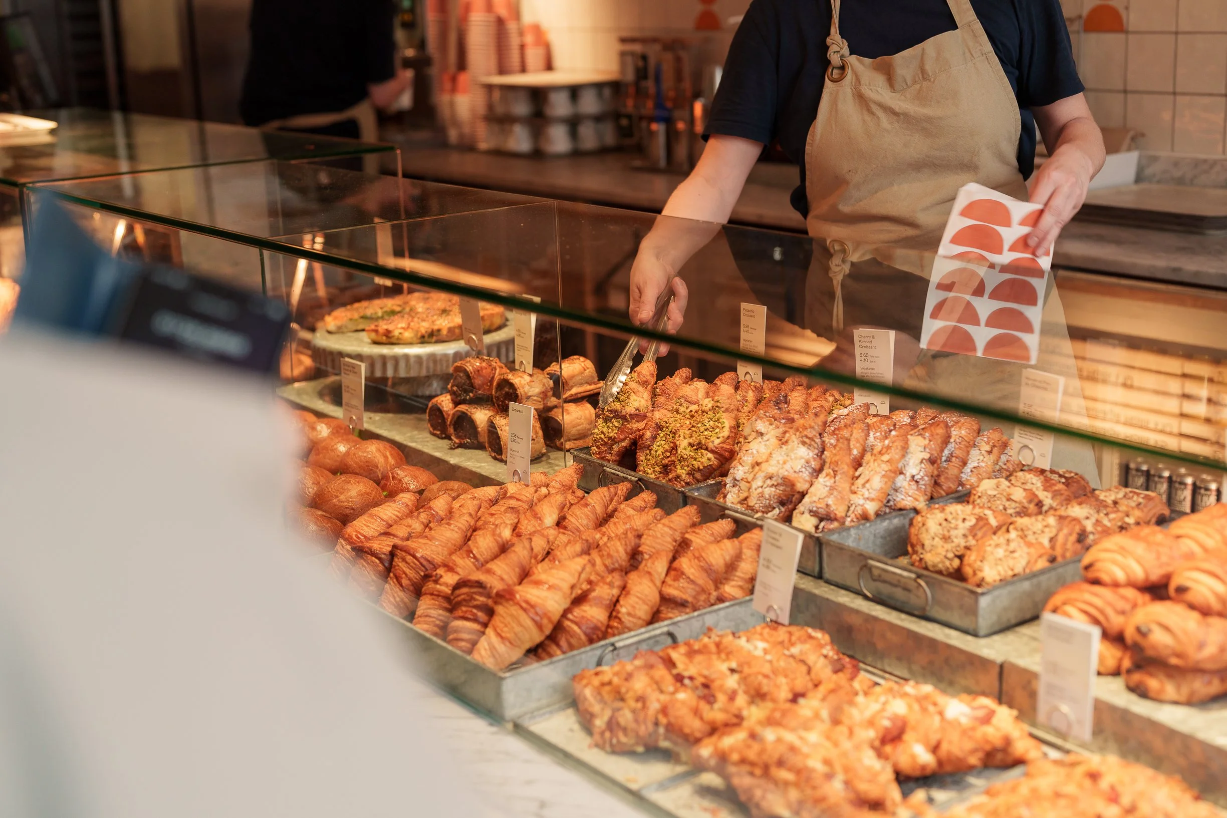 Display case with various baked goods including croissants, pastries, and bread at a bakery counter, with a staff member arranging treats.