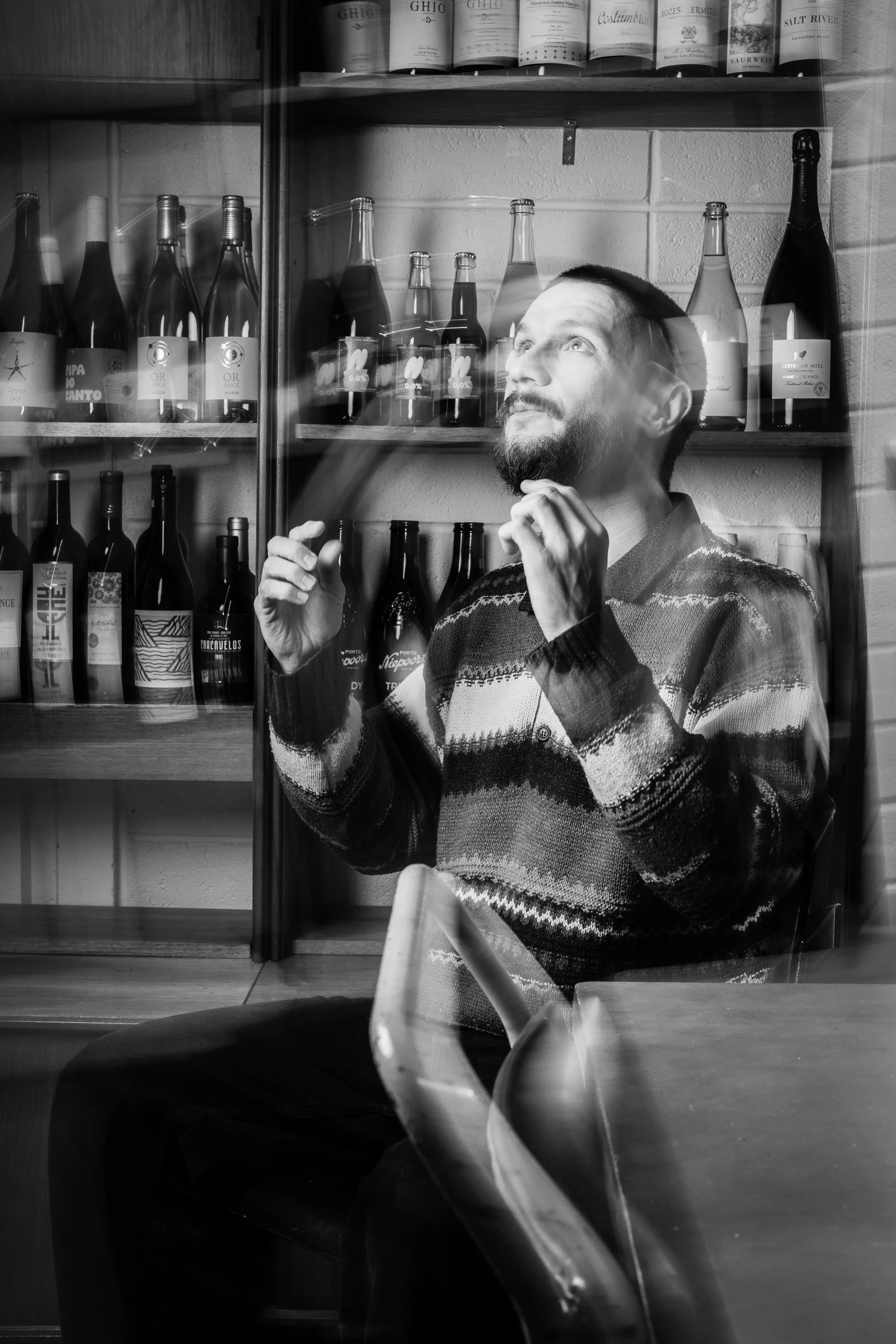 A man with a beard wearing a patterned sweater sitting indoors behind a glass window, with wine bottles on shelves in the background.