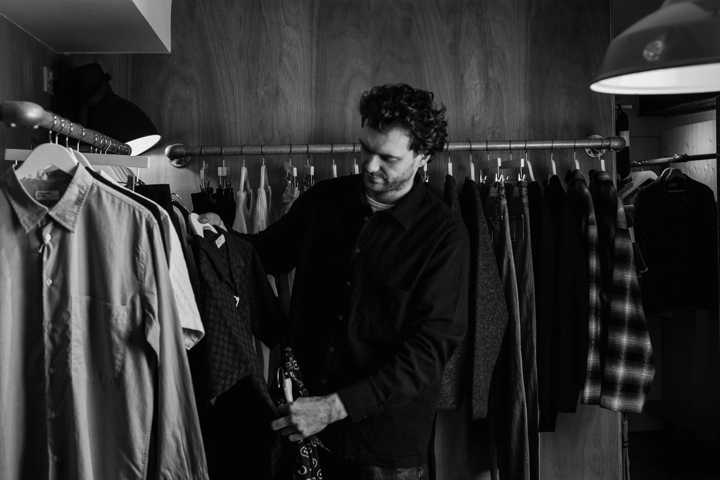 A man with curly hair shopping for clothes in a store, examining a dark-colored jacket among other garments hanging on a clothing rack.