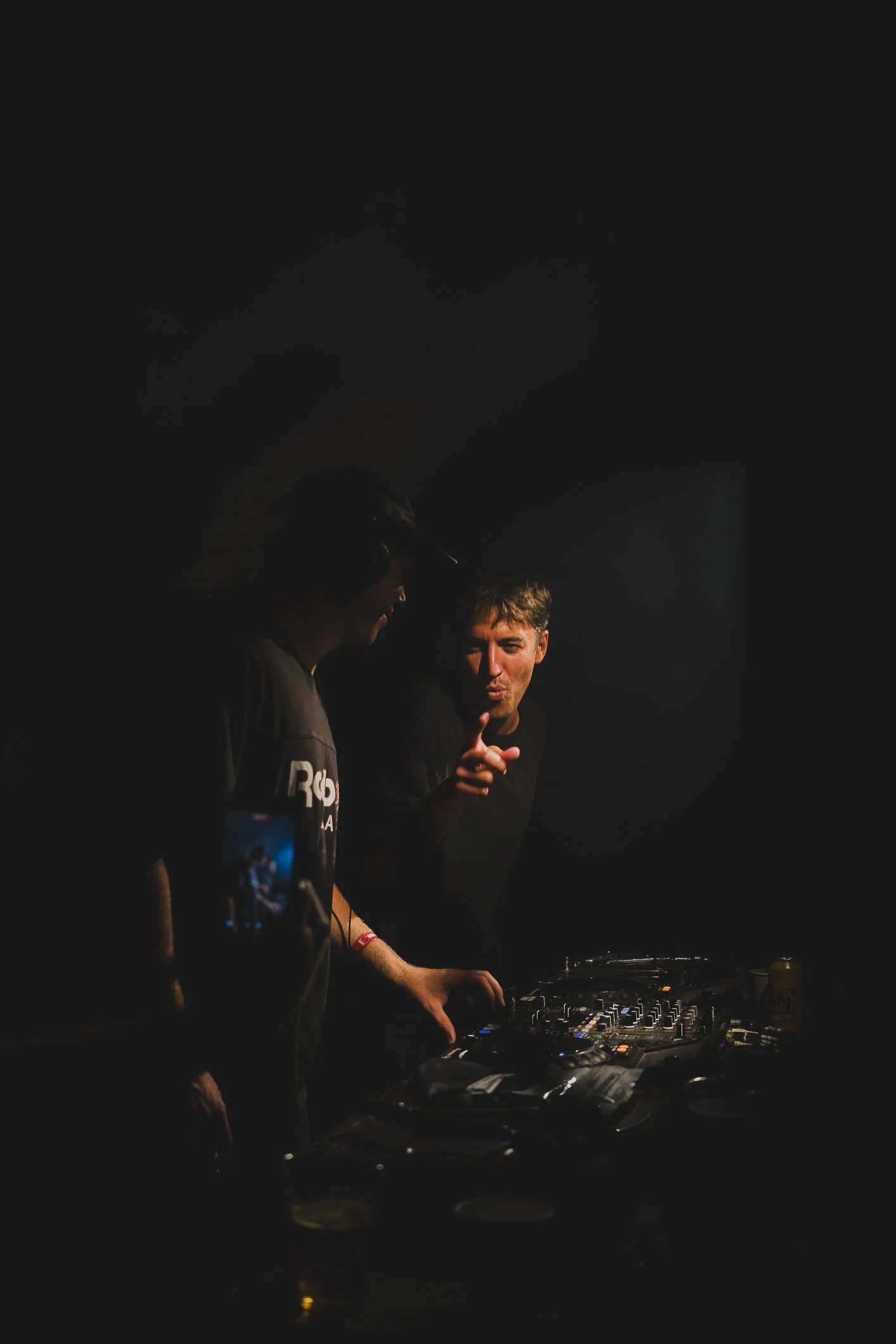 Two DJs, one in a black shirt and the other in a black cap, are performing at a DJ booth in a dark setting. The man in the cap is listening, while the other is pointing and speaking. DJ equipment and a beverage can are visible on the table.