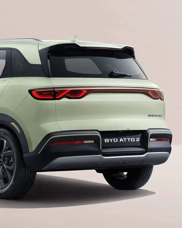 Zbliżenie na tył elektrycznego SUV-a BYD Atto 2 w jasnozielonym lakierze, prezentujące tylną szybę ze spoilerem, charakterystyczny pas świetlny LED oraz tylne koła na jasnym tle.