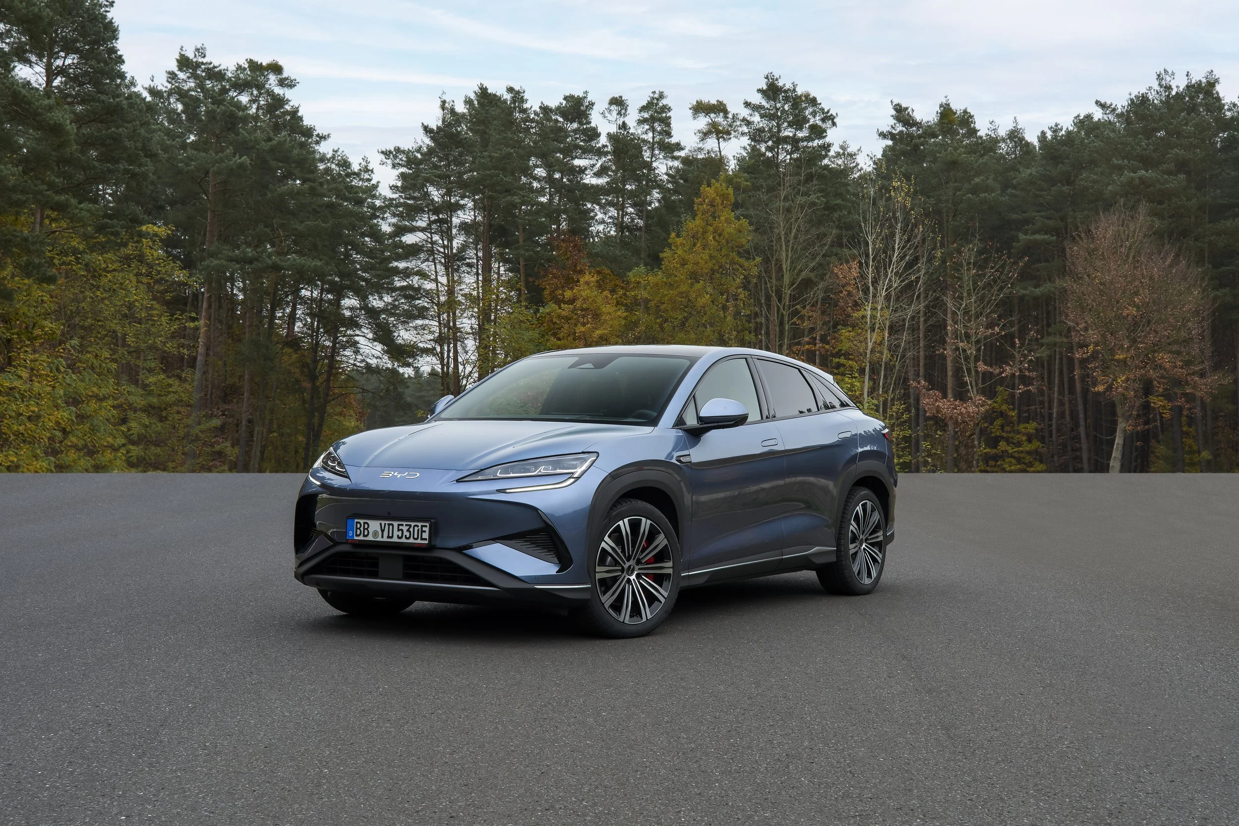 Niebieski elektryczny SUV BYD Sealion 7 zaparkowany na asfalcie na tle gęstego lasu, ukazujący opływową sylwetkę i wąskie reflektory, prezentowany przy salonie BYD Garcarek.