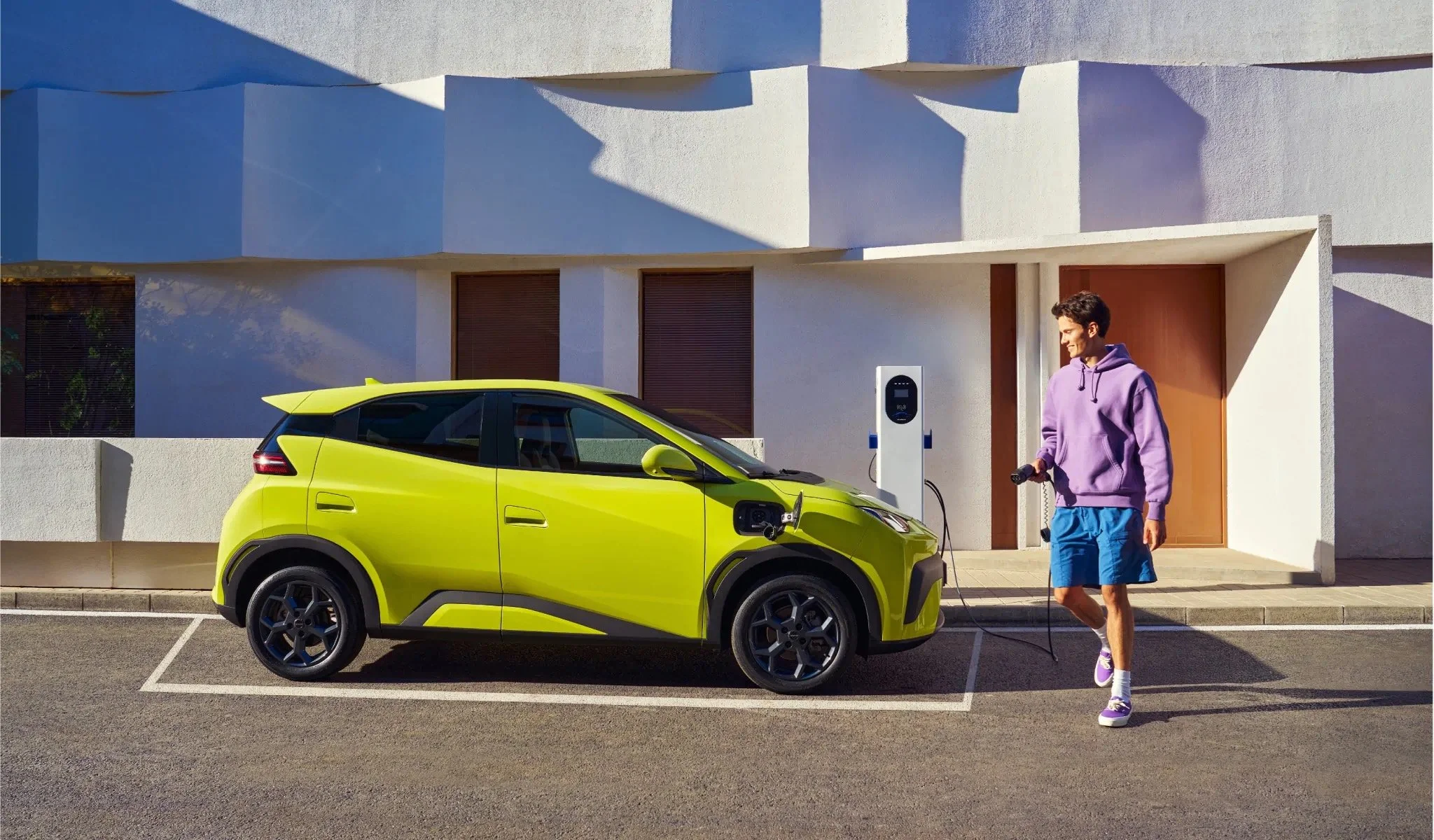 Bok elektrycznego samochodu BYD Dolphin Mini w limonkowym kolorze stojącego przy nowoczesnym budynku z otwartym portem ładowania i widoczną w tle stacją wallbox.