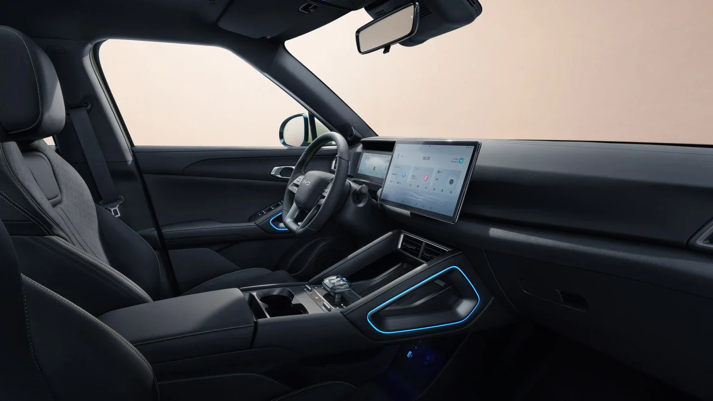 Wnętrze modelu BYD Atto 2 od strony pasażera, przedstawiające deskę rozdzielczą z dużym ekranem dotykowym, kierownicę, konsolę środkową z niebieskim podświetleniem ambientowym oraz lusterko wsteczne na jasnym tle.