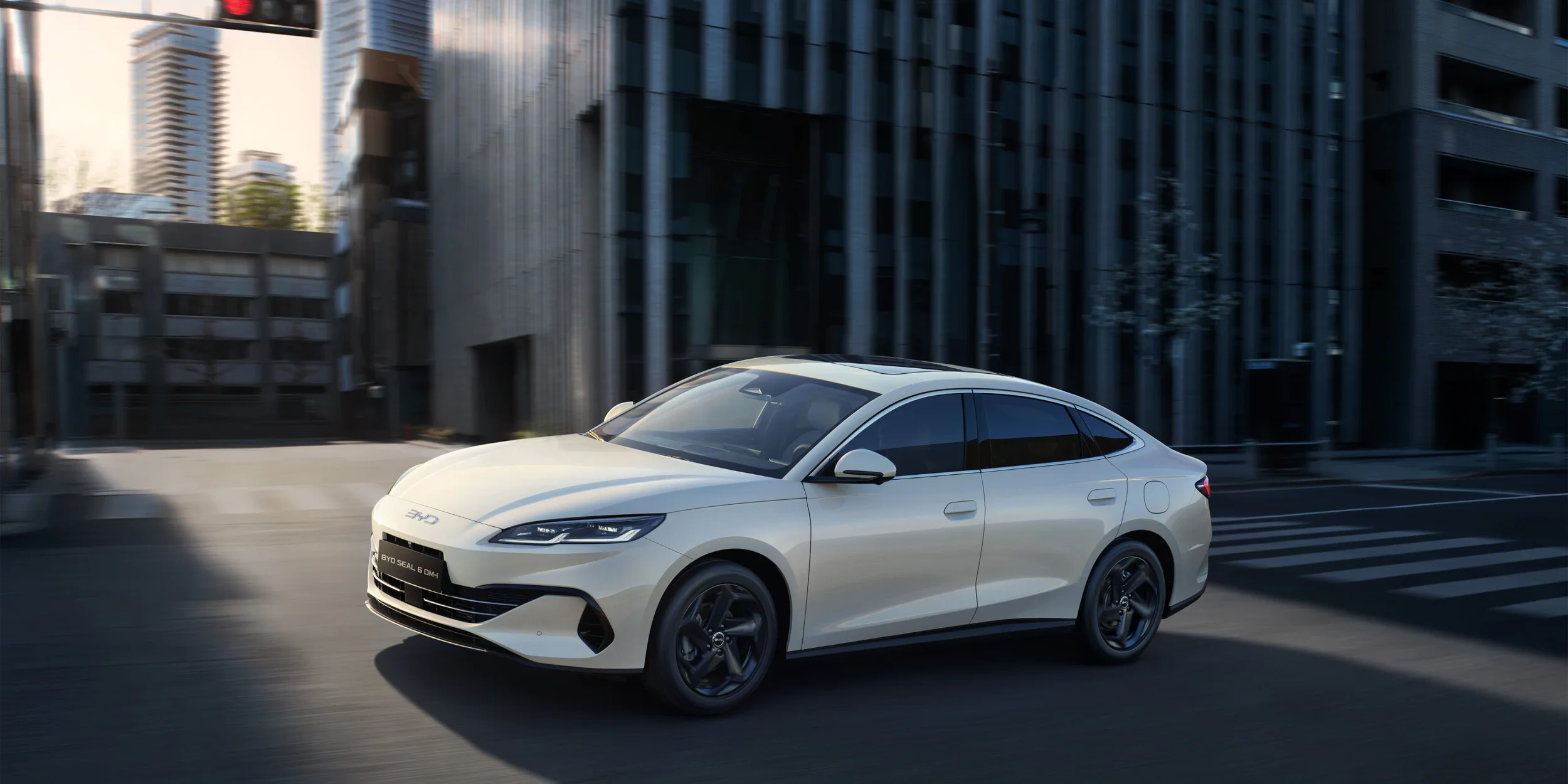 Biały hybrydowy sedan BYD Seal 6 DM-i w dynamicznym ruchu miejskim na tle rozmytych nowoczesnych wieżowców, sfotografowany pod kątem trzech czwartych z przodu.