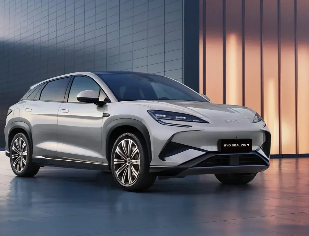 Srebrny elektryczny SUV BYD Sealion 7 zaparkowany na nowoczesnym placu obok wolnostojącej ładowarki samochodowej, na tle minimalistycznego budynku o ciemnej, kafelkowej elewacji.