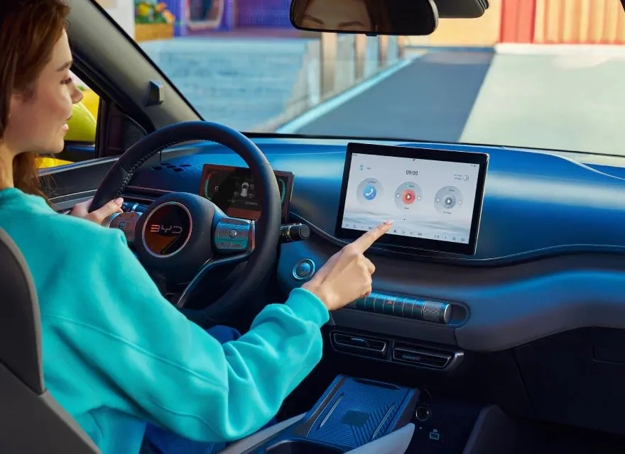 Wnętrze modelu BYD Dolphin Surf z perspektywy kierowcy, przedstawiające dłoń obsługującą centralny ekran dotykowy, trójramienną kierownicę z logo BYD, cyfrowy wyświetlacz wskaźników oraz widok przez przednią szybę.