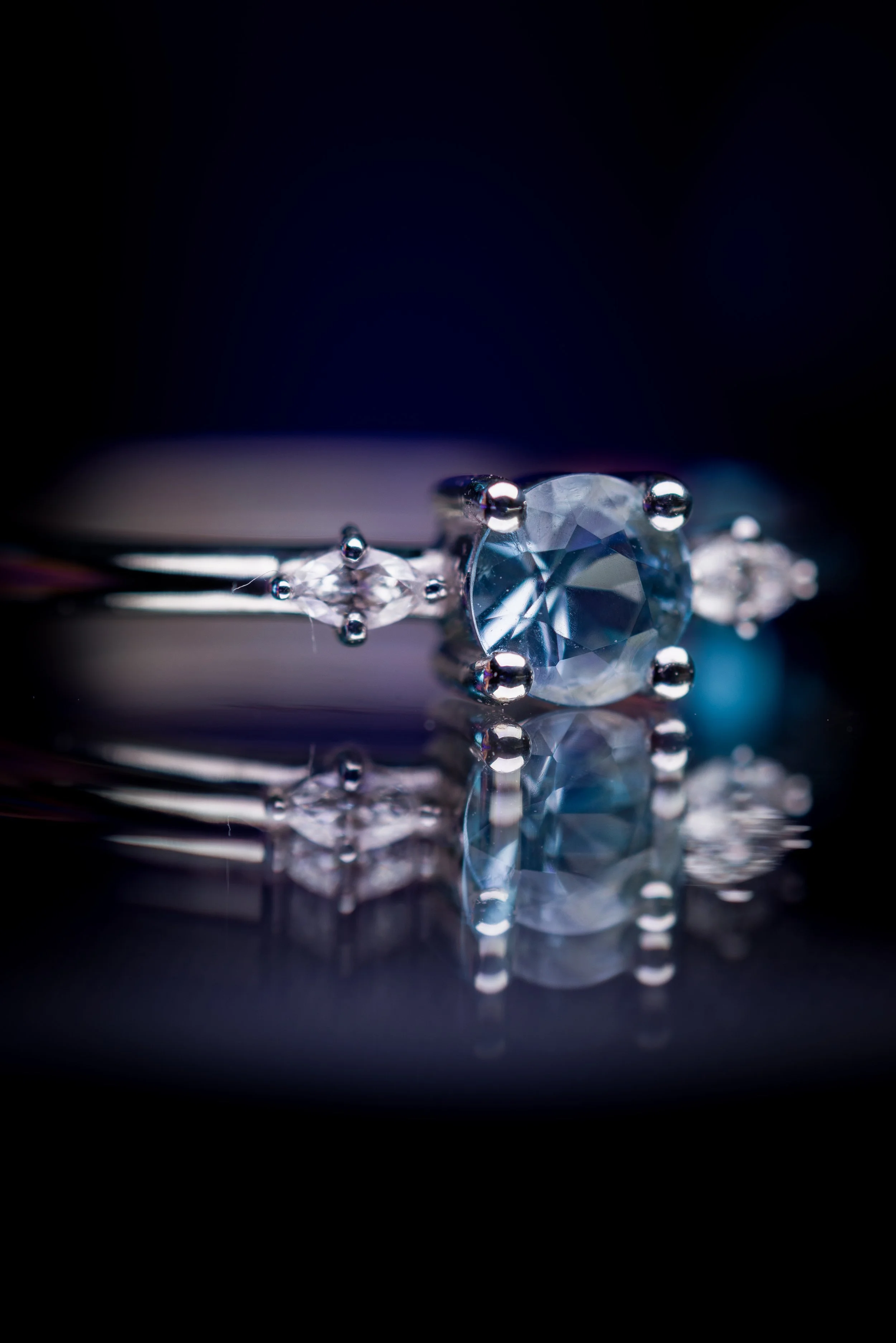 Bague en or blanc avec une aigue marine et des diamants navettesant central rond entouré de deux petits diamants, posée sur une surface noire reflétant la bague.