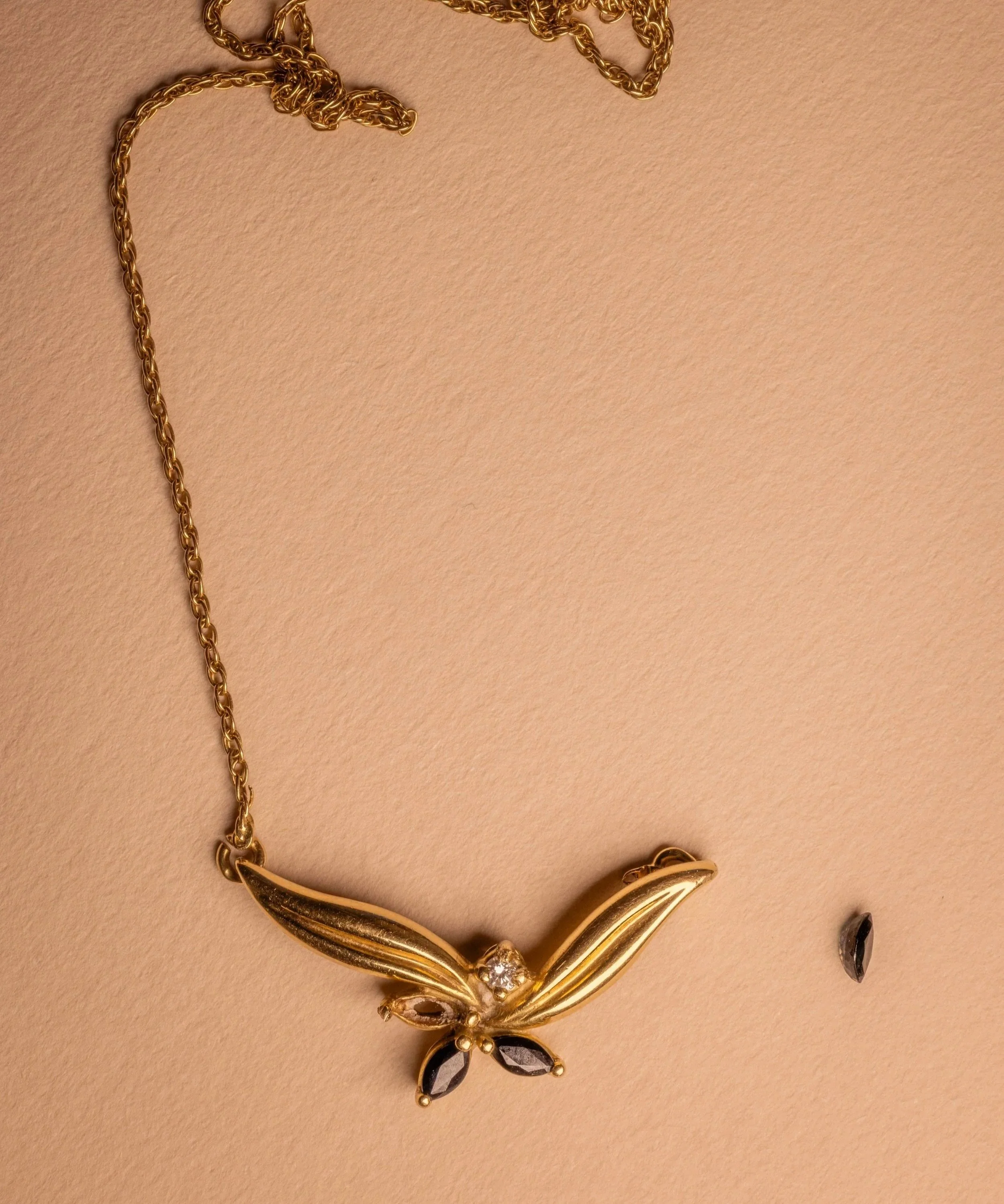 Collier avant d'être transformé en or avec un pendentif en forme de papillon orné de saphirs bleus et un diamant, posé sur un fond beige avec un saphir bleu desserti à côté.