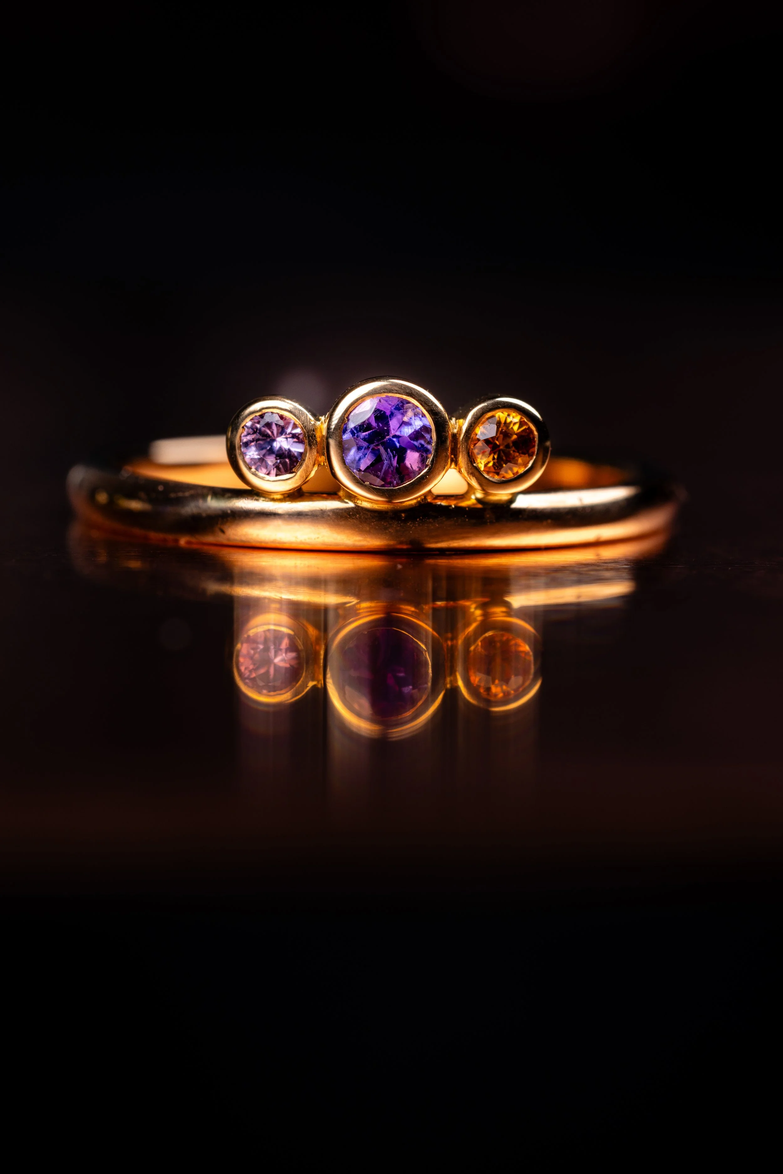 Bague en or avec trois pierres précieuses tanzanite, saphir rose et saphir orange, reflet sur surface noire.