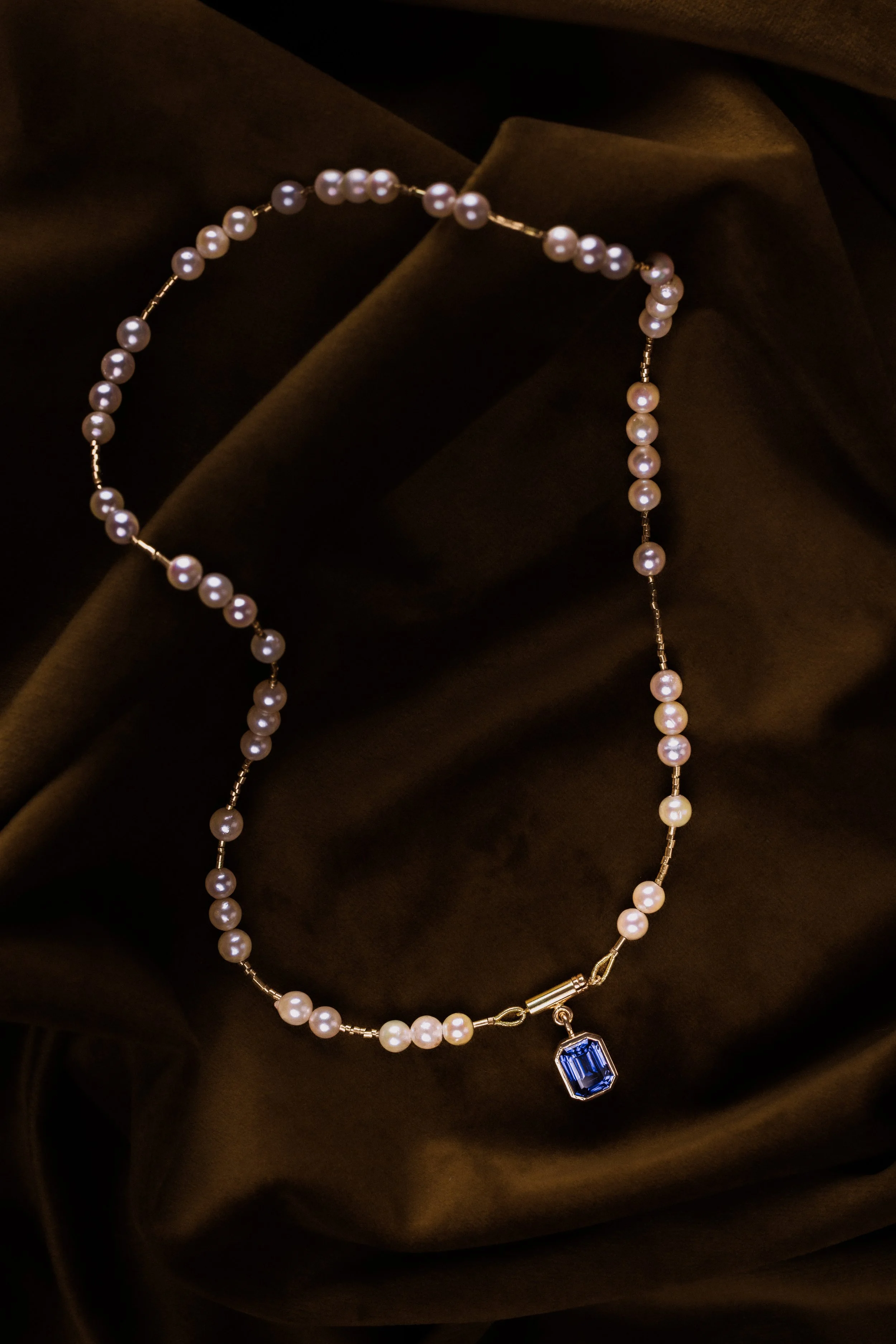 Collier de perles de culture avec pendentif en saphir bleuté, posé sur un tissu sombre.