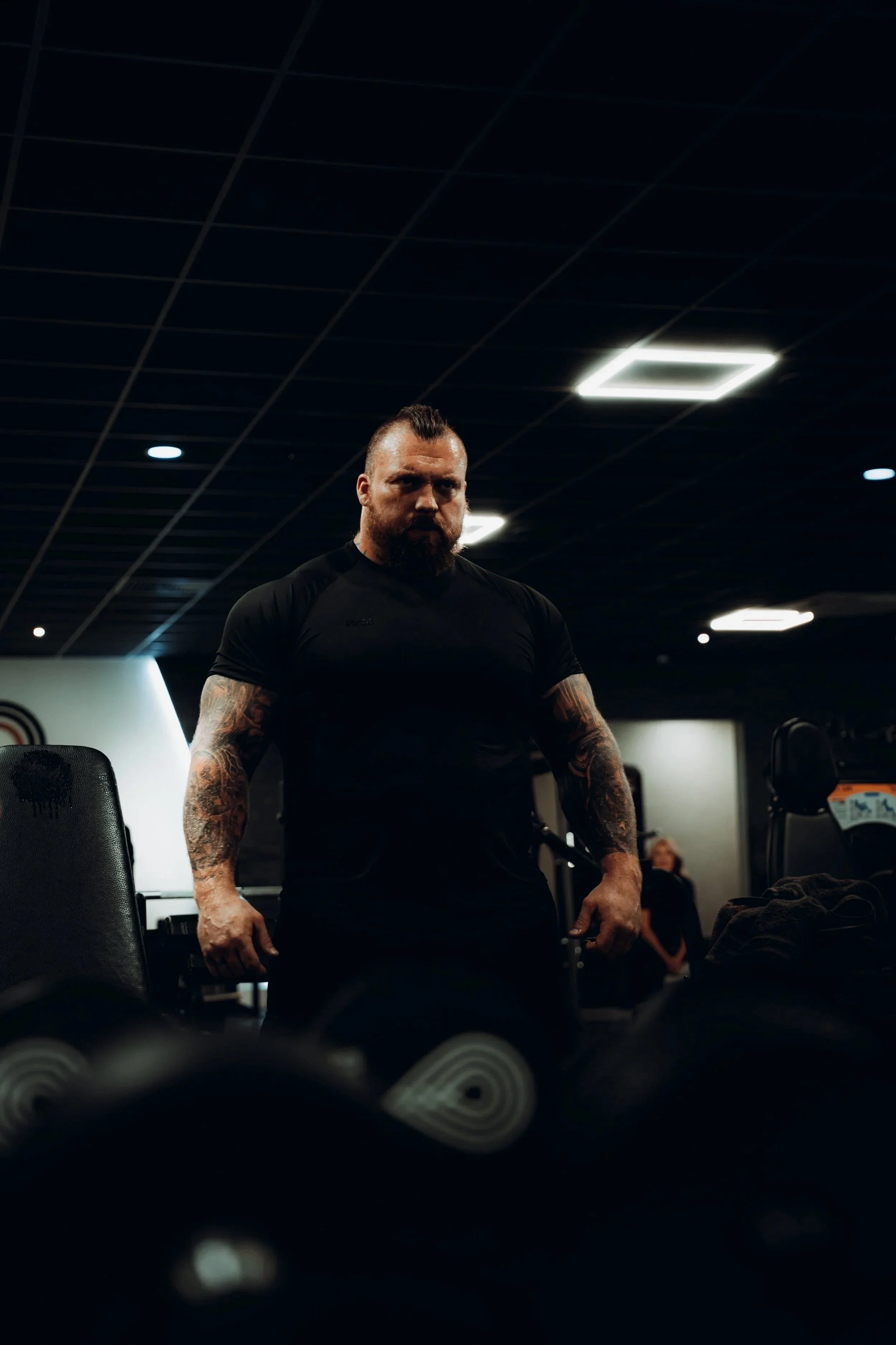 A muscular man with tattoos looks intensely while standing in a gym filled with workout equipment and dim lighting.