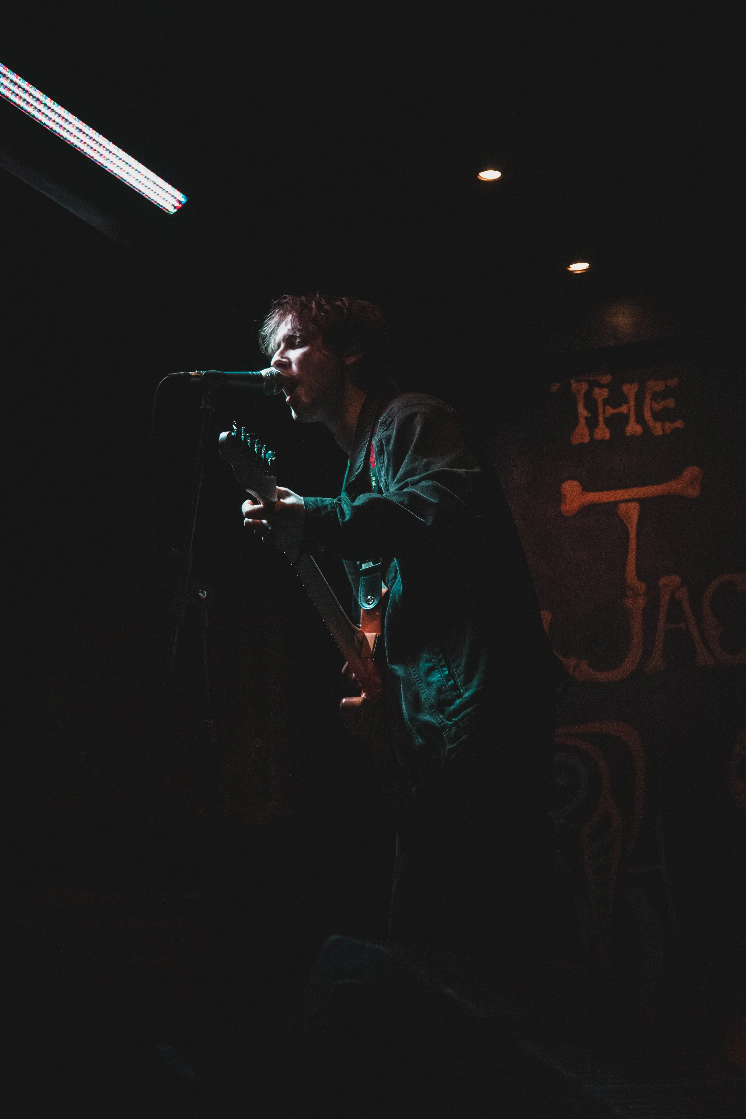 A musician performing on stage with a guitar and microphone, in a dark venue with a logo or name in the background.