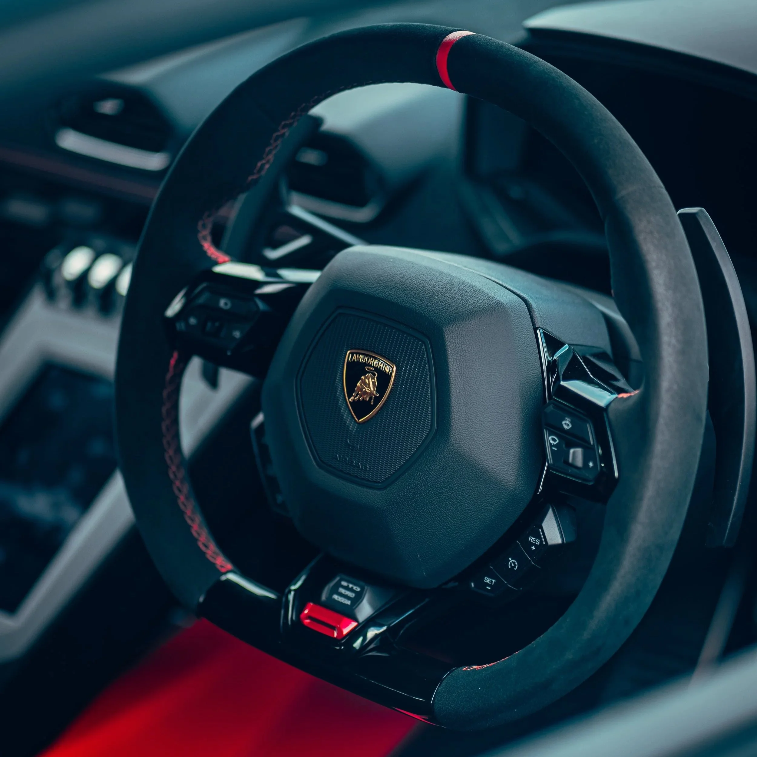 Close-up of a black Lamborghini car steering wheel with the Lamborghini logo in the center, highlighted in gold, featuring various control buttons.