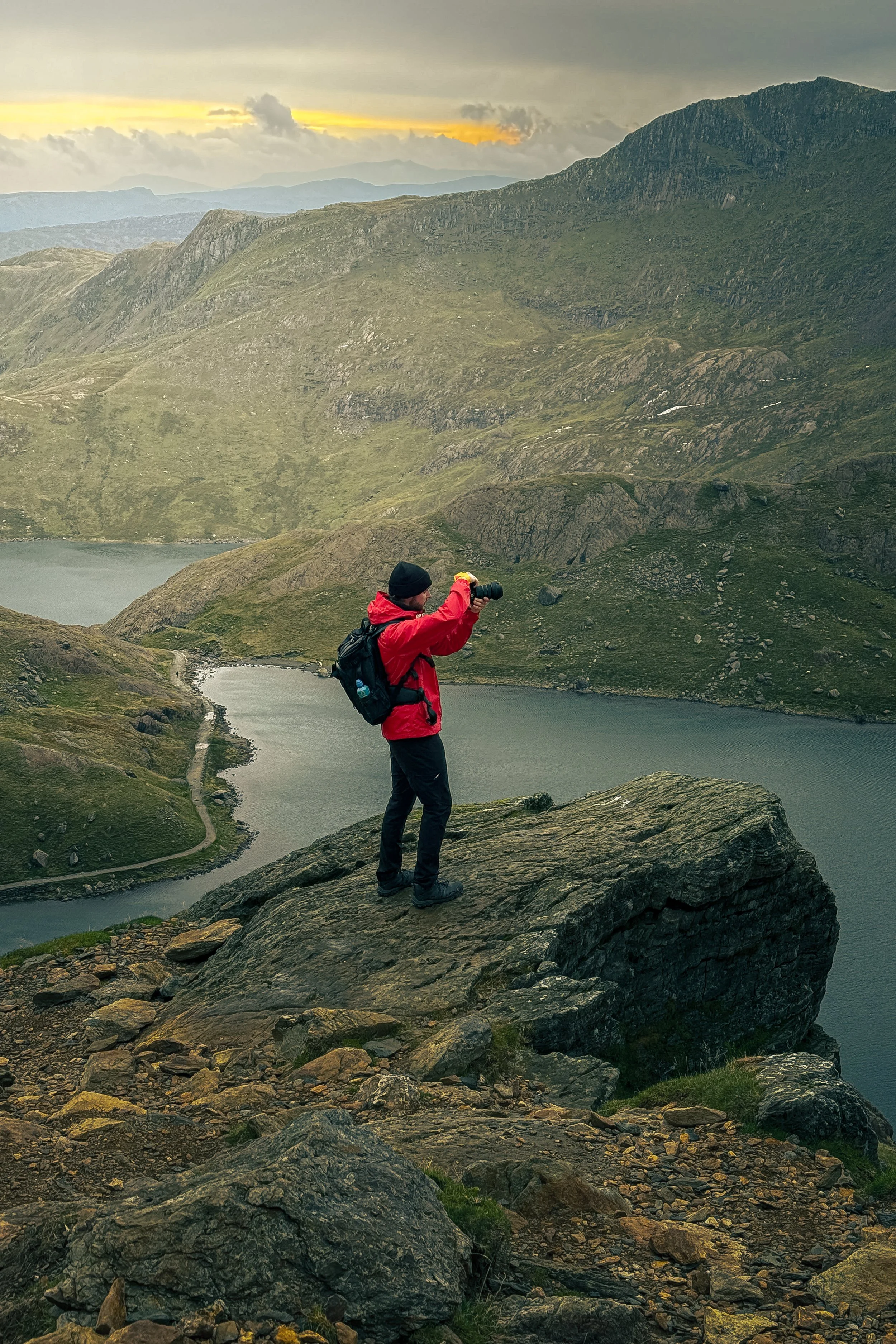 A hiker in a red jacket and black hat standing on a large rock, taking a photo of a scenic mountain landscape with a lake and cloudy sky during sunset or sunrise.