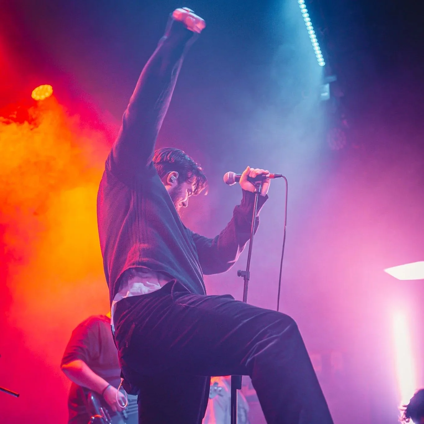 A male singer performing on stage, holding a microphone and raising one arm, with colorful stage lighting and a band member in the background.