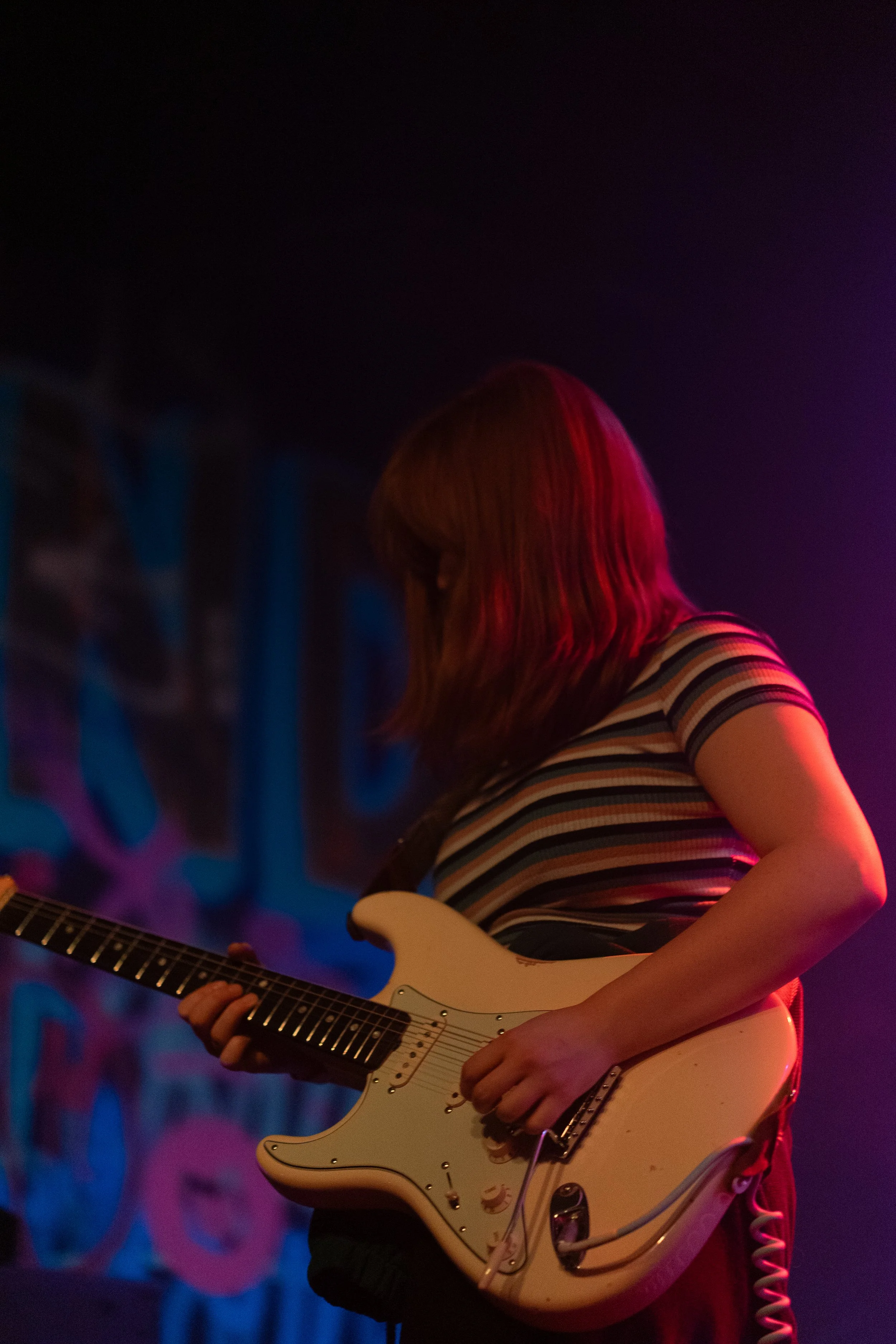 A woman with shoulder-length hair playing an electric guitar on stage with colorful lighting.