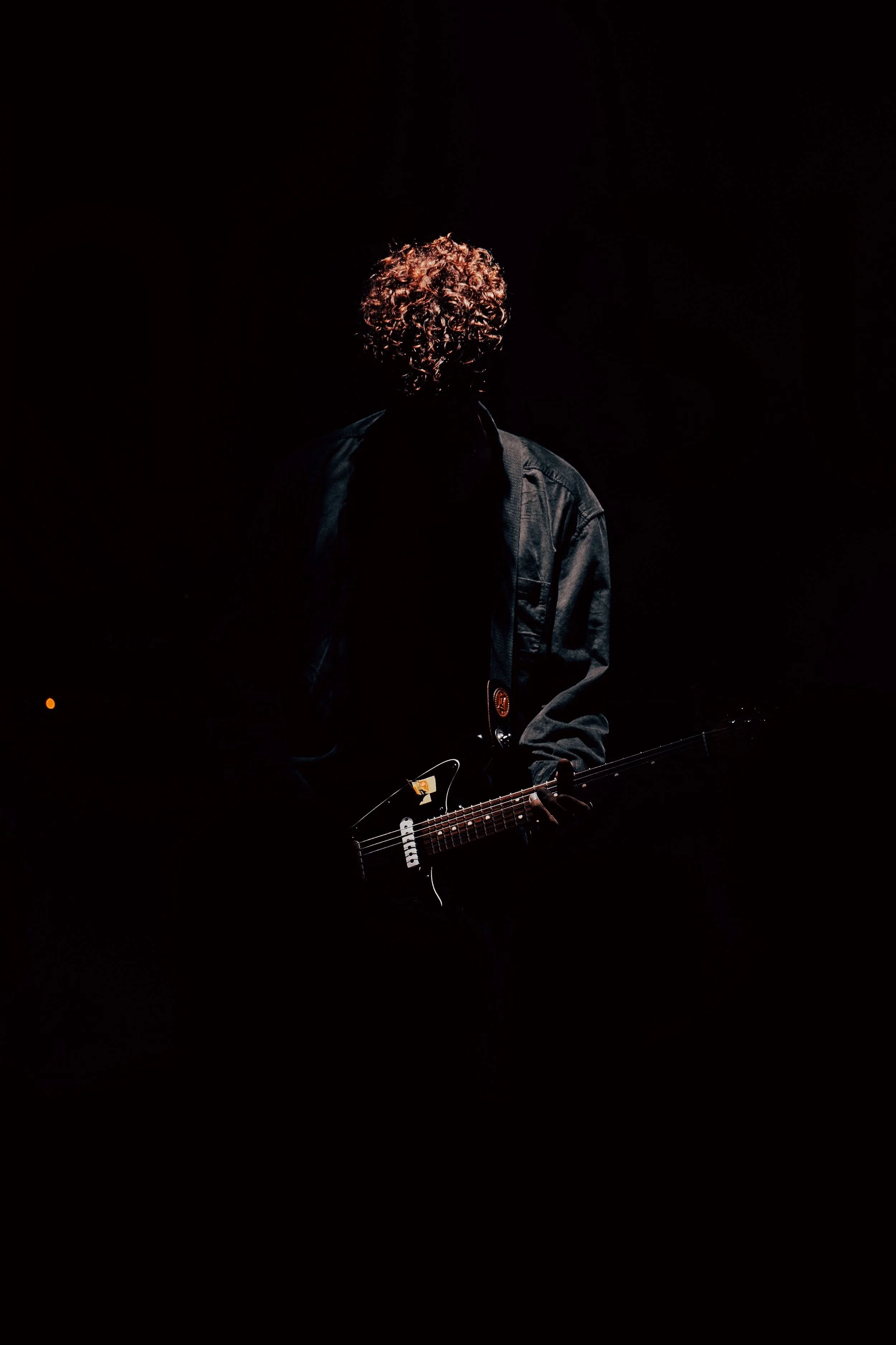 Person with curly hair playing guitar in a dark setting with spotlighting.