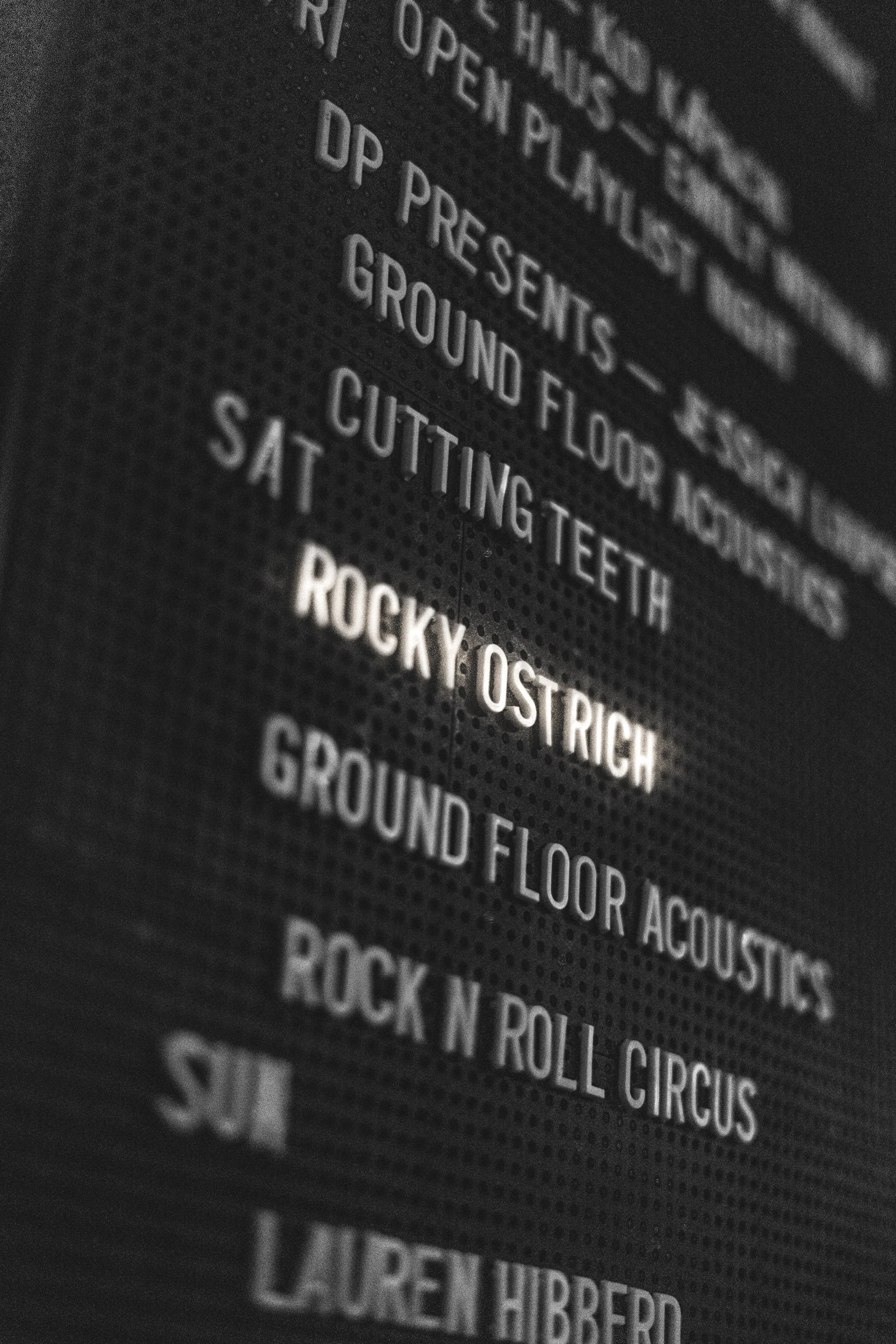 Close-up view of a digital display with a list of musical acts and songs, including Rocky O'Strich and the song 'Sat.'