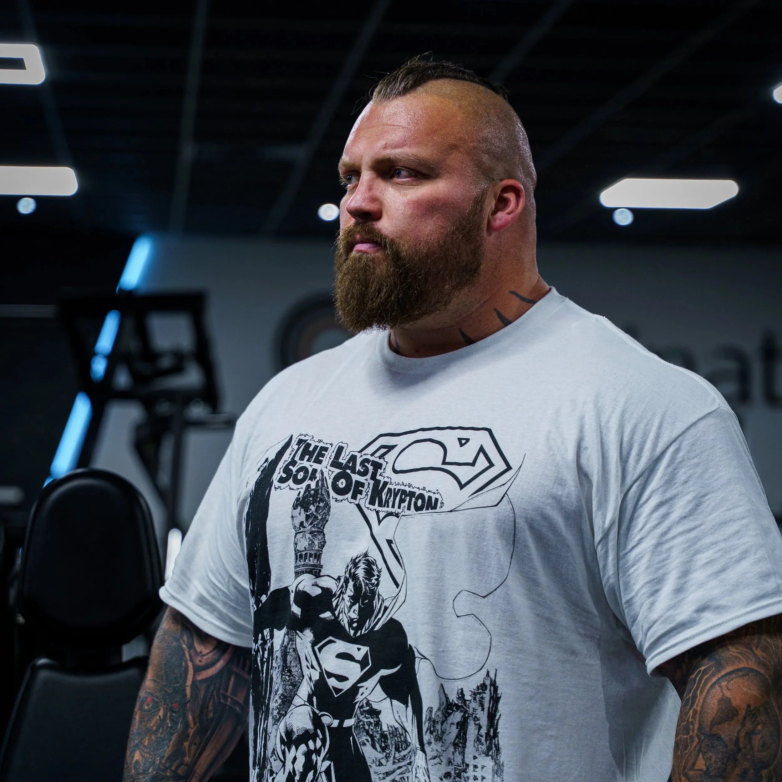 A muscular man with a beard and slicked-back hair in a gym, wearing a white T-shirt featuring comic book Superman artwork and the text "The Last Son of Krypton."