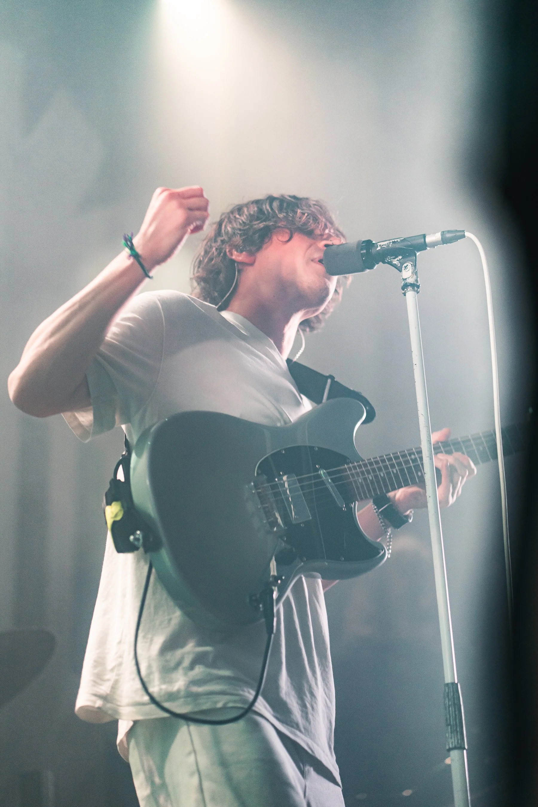 Musician singing into microphone while playing an electric guitar on stage with bright light.