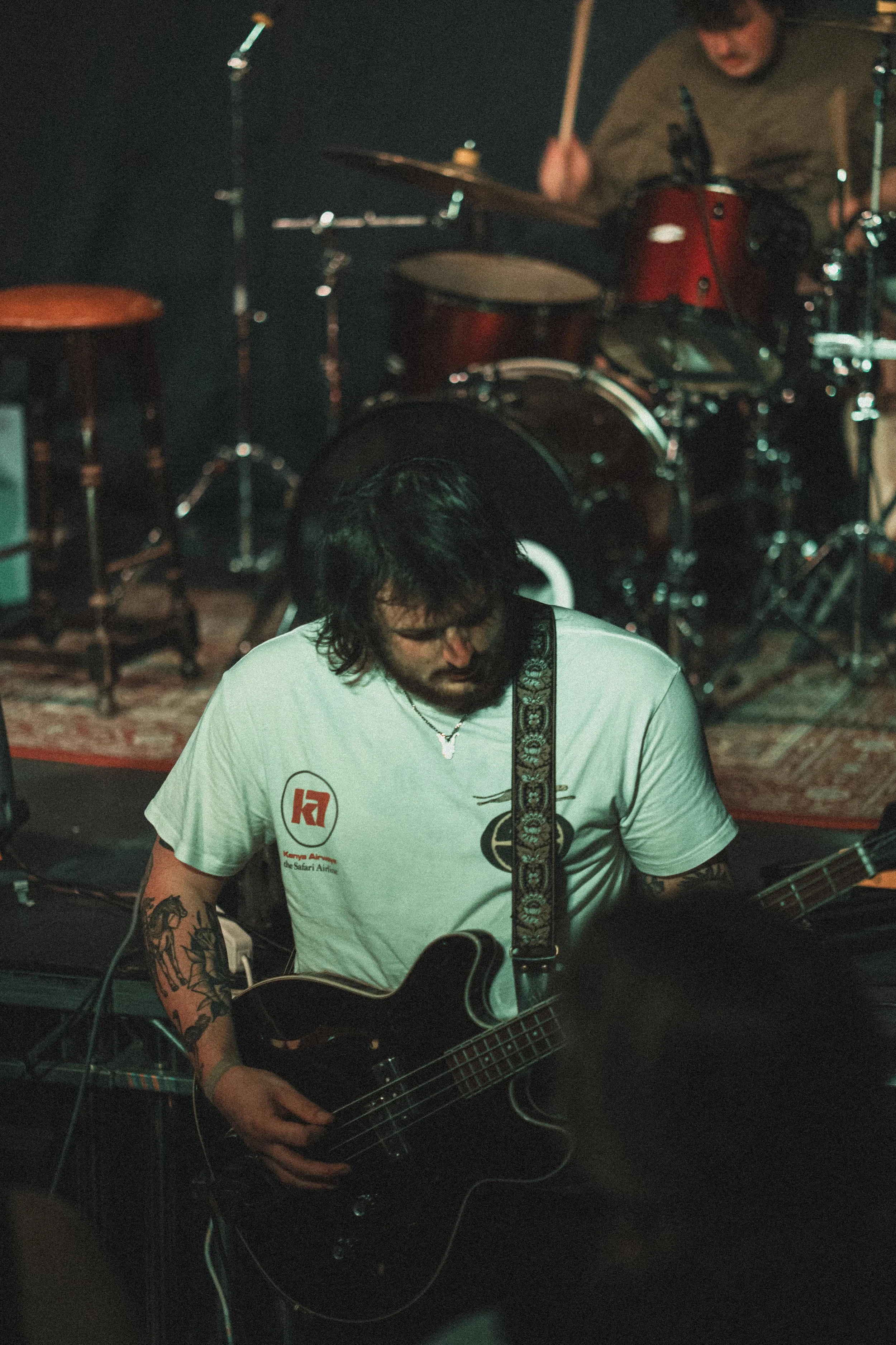 A musician with dark hair and glasses playing an electric bass guitar on stage, with a drummer in the background, in a dimly lit venue.