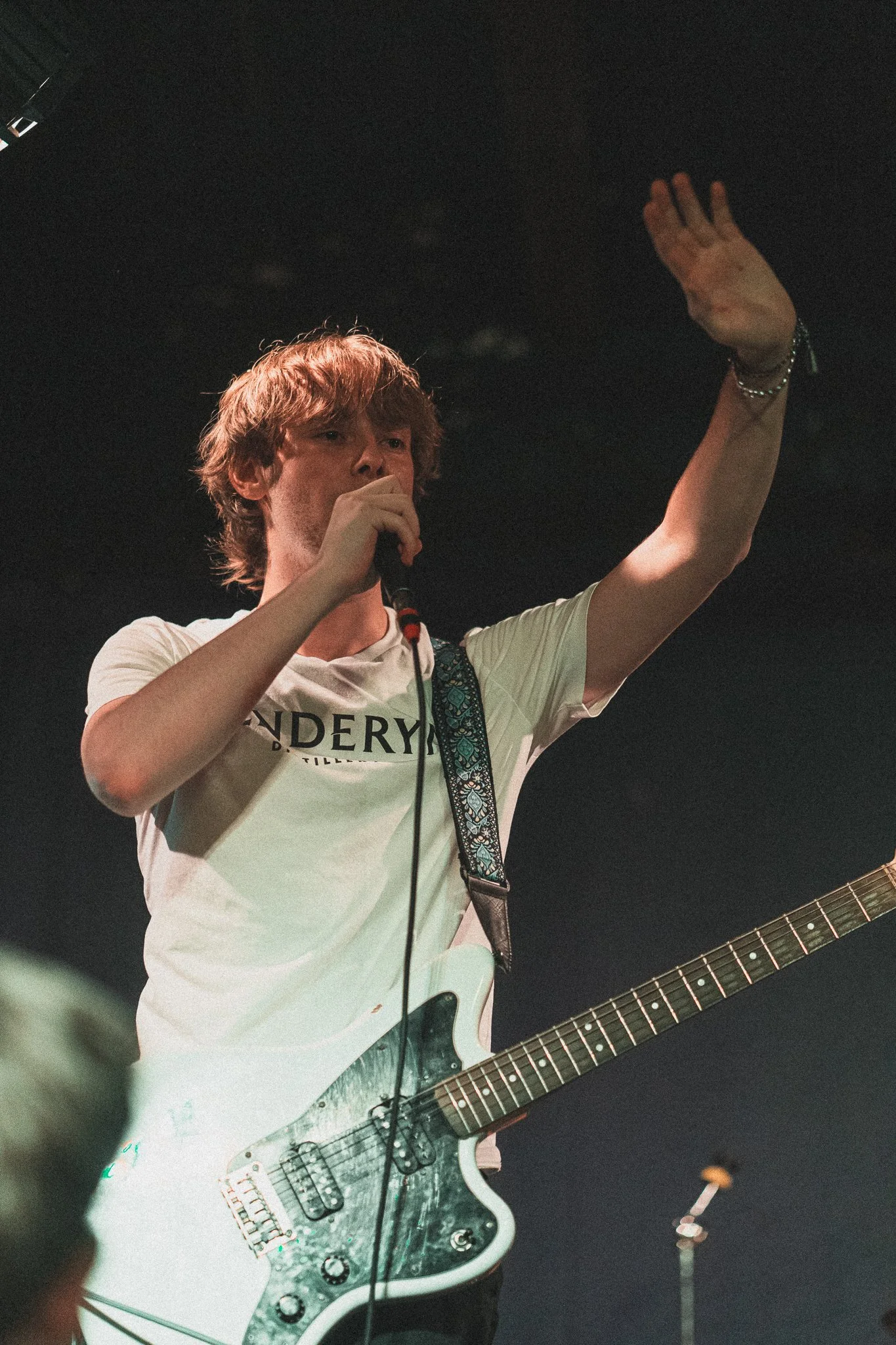 Young man with brown hair singing into a microphone while holding an electric guitar, wearing a white T-shirt and a bracelet, on stage under dark lighting.