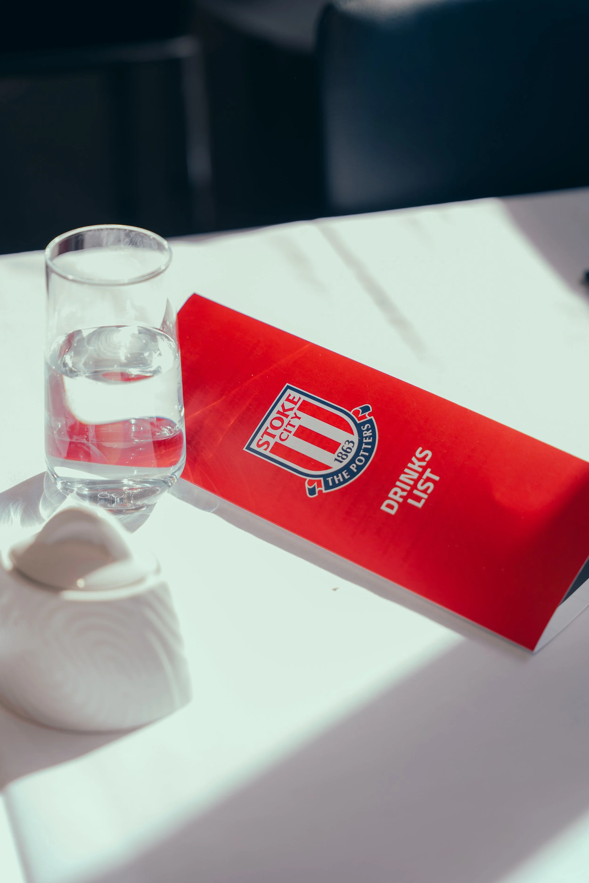 A clear glass of water and a red drinks list booklet on a white table with sunlight and shadows.
