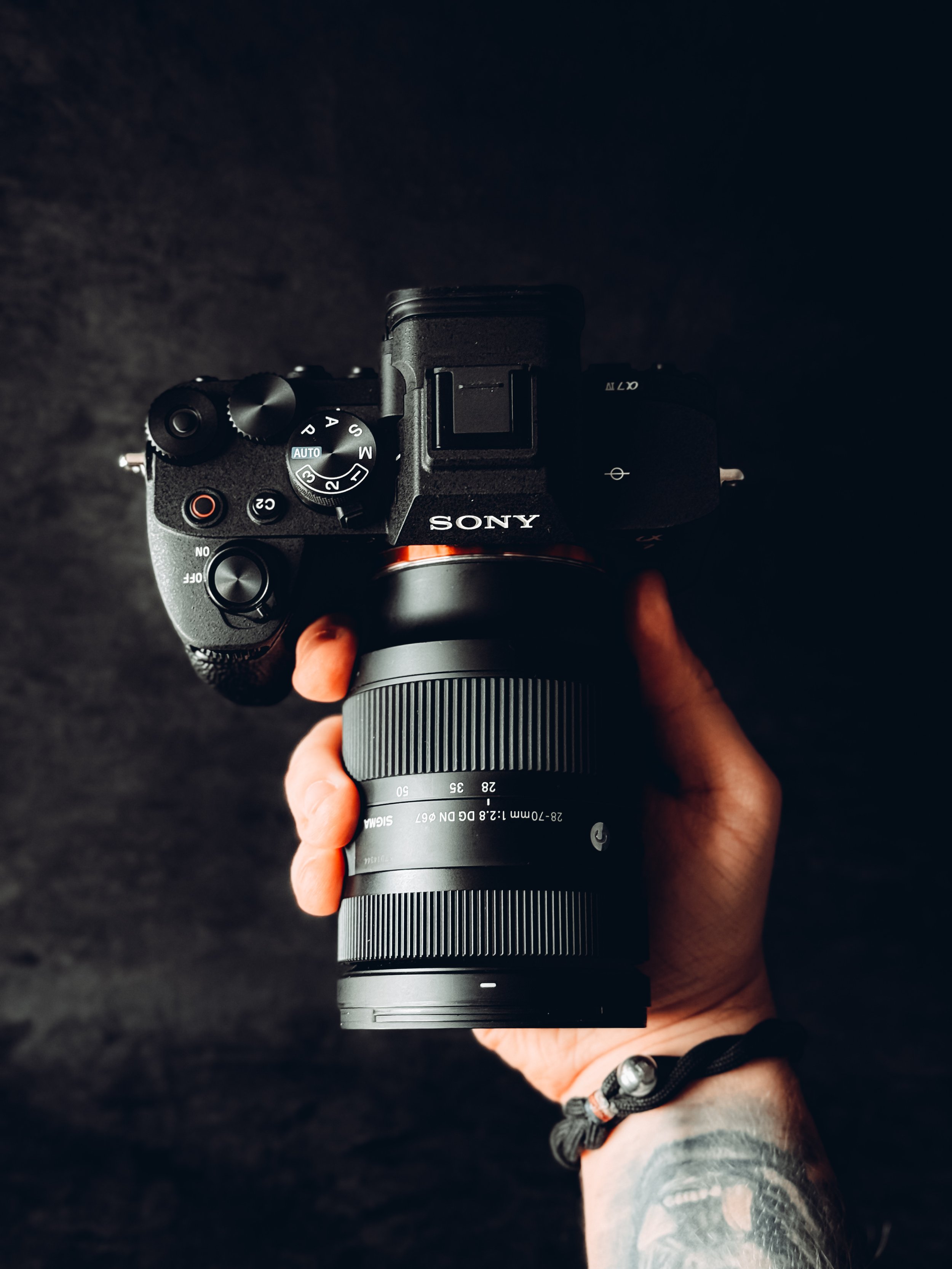 Hand holding a Sony mirrorless camera with a zoom lens against a dark background.
